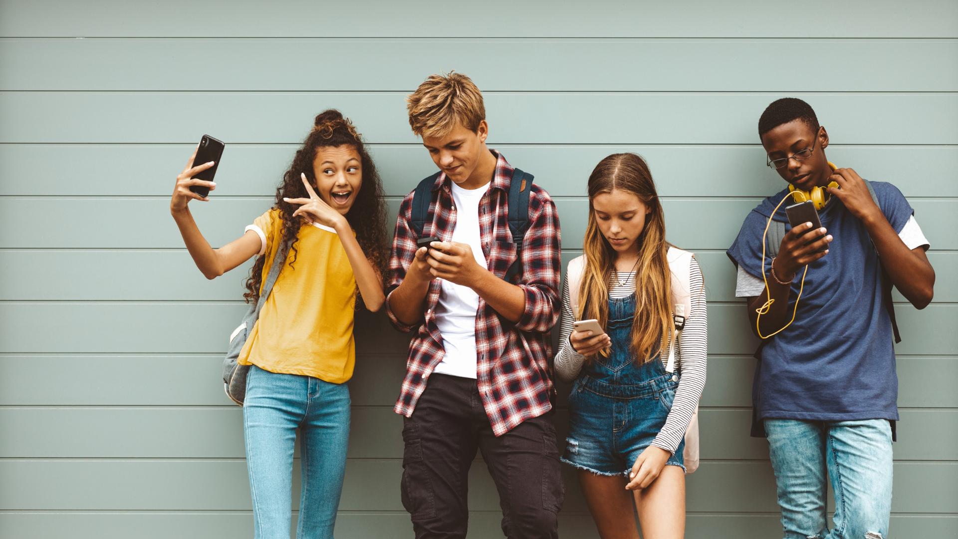 Four teenagers standing against a light gray wall, each looking at their smartphones. The girl on the left, wearing a yellow top and jeans, is smiling and holding her phone up, excited. The boy next to her, in a plaid shirt and backpack, is focused on his phone. The girl beside him, in a striped top and denim shorts, is reading her phone with a neutral expression. The boy on the far right, wearing glasses, a blue shirt, headphones, and ripped jeans, is also looking at his phone.