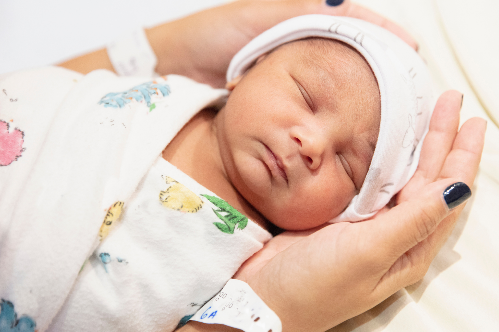 A sleeping newborn baby with a hospital wristband, resting in a person's hand, wearing a hospital cap and a patterned blanket.