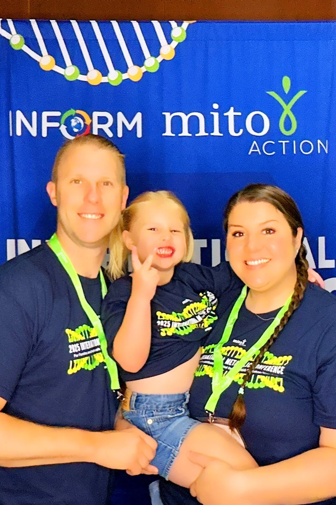 A smiling family of three posing in front of a blue 'INFORM mito ACTION' banner, all wearing matching navy blue shirts with yellow and white text and green lanyards.