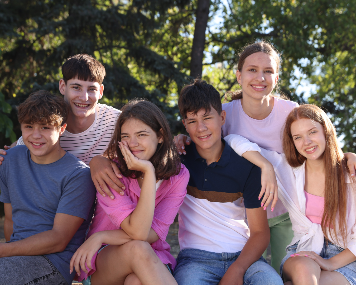 Group of seven teenagers smiling and sitting outdoors with trees in the background.