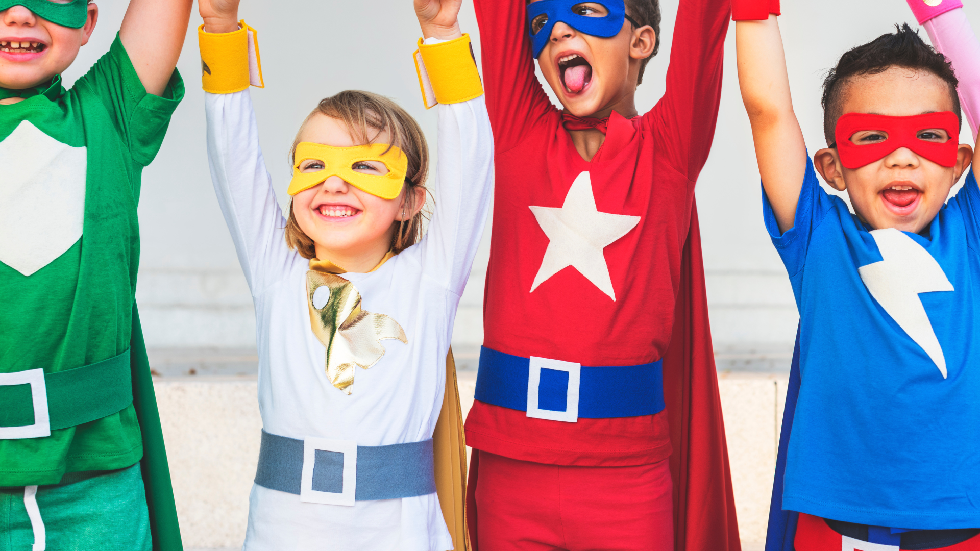 Four children dressed in superhero costumes, smiling and raising their arms.