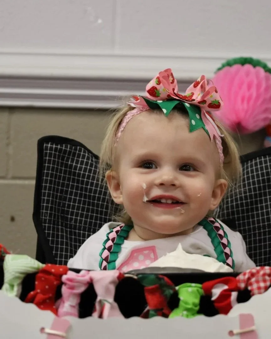 A young girl with blonde hair, wearing a pink headband with a strawberry-themed bow and a birthday bib, sitting in a high chair with remnants of cake around her mouth, celebrating her birthday.