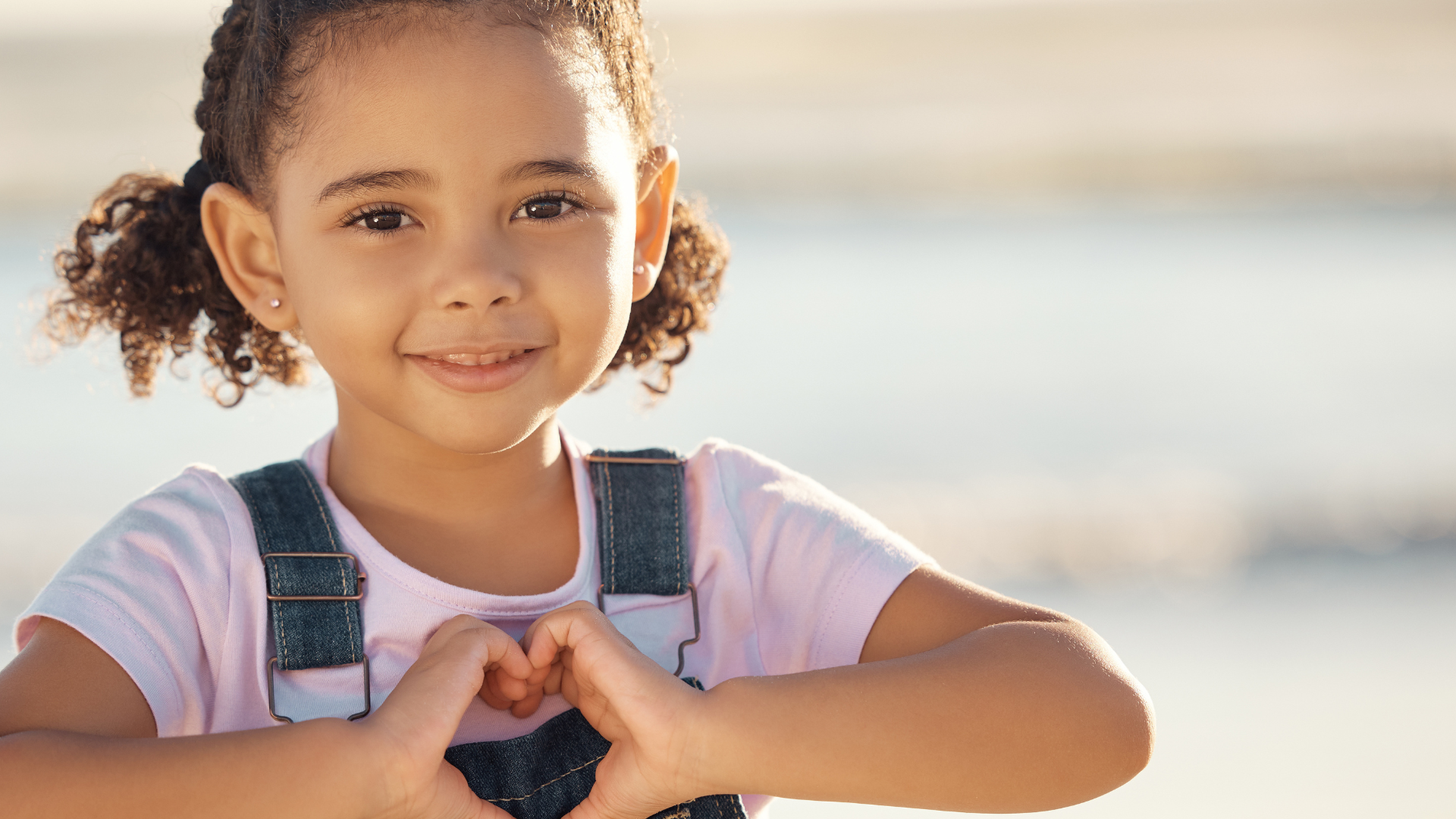 A young girl with curly hair making a heart shape with her hands outdoors near water.