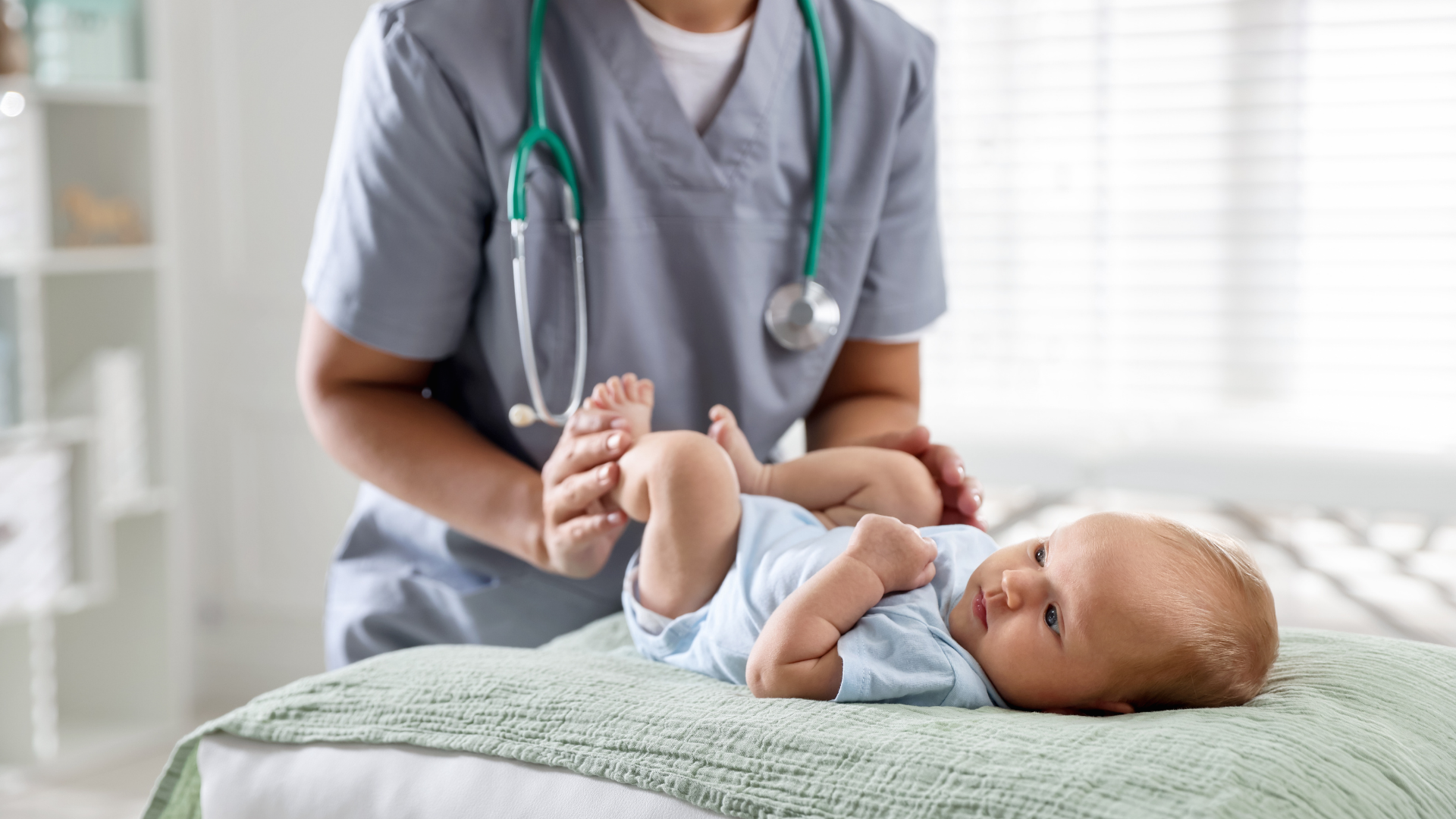 A healthcare professional examining a baby on a hospital bed, with a stethoscope around their neck, in a bright medical room.