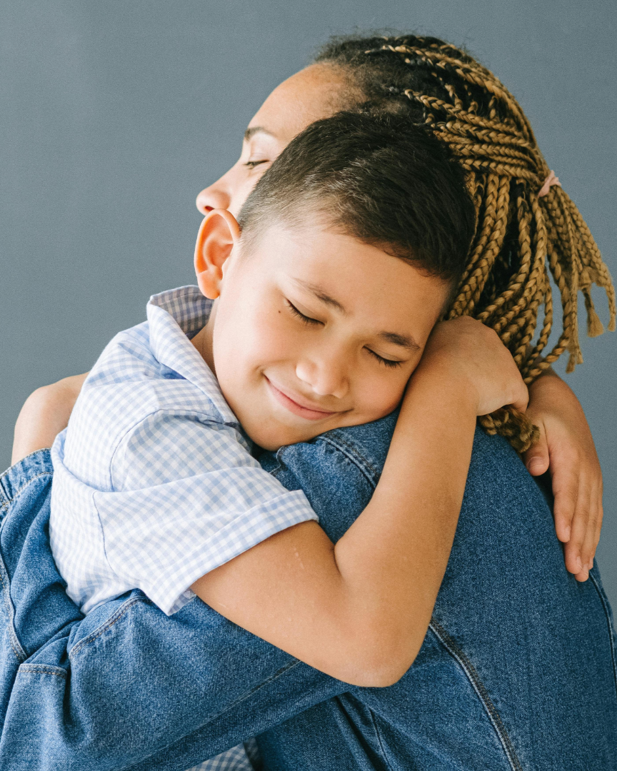 A young boy with short dark hair and a checked shirt hugging an adult with braided hair, both with eyes closed, showing affection and comfort.