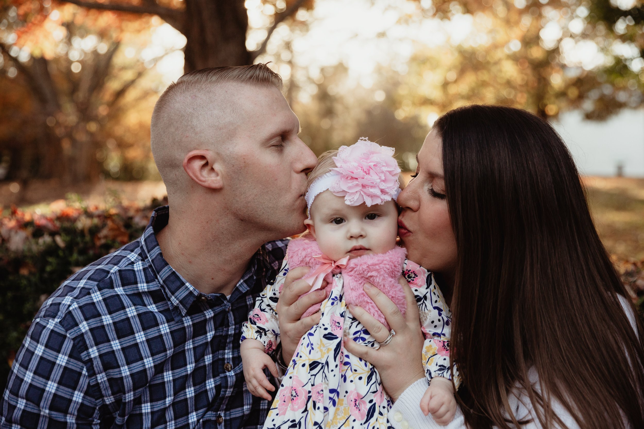 A family of three, including a man, woman, and a baby girl, outdoors during fall. Both adults are kissing the baby on the cheeks. The baby is wearing a pink headband with a large flower and a floral dress, held by the woman.