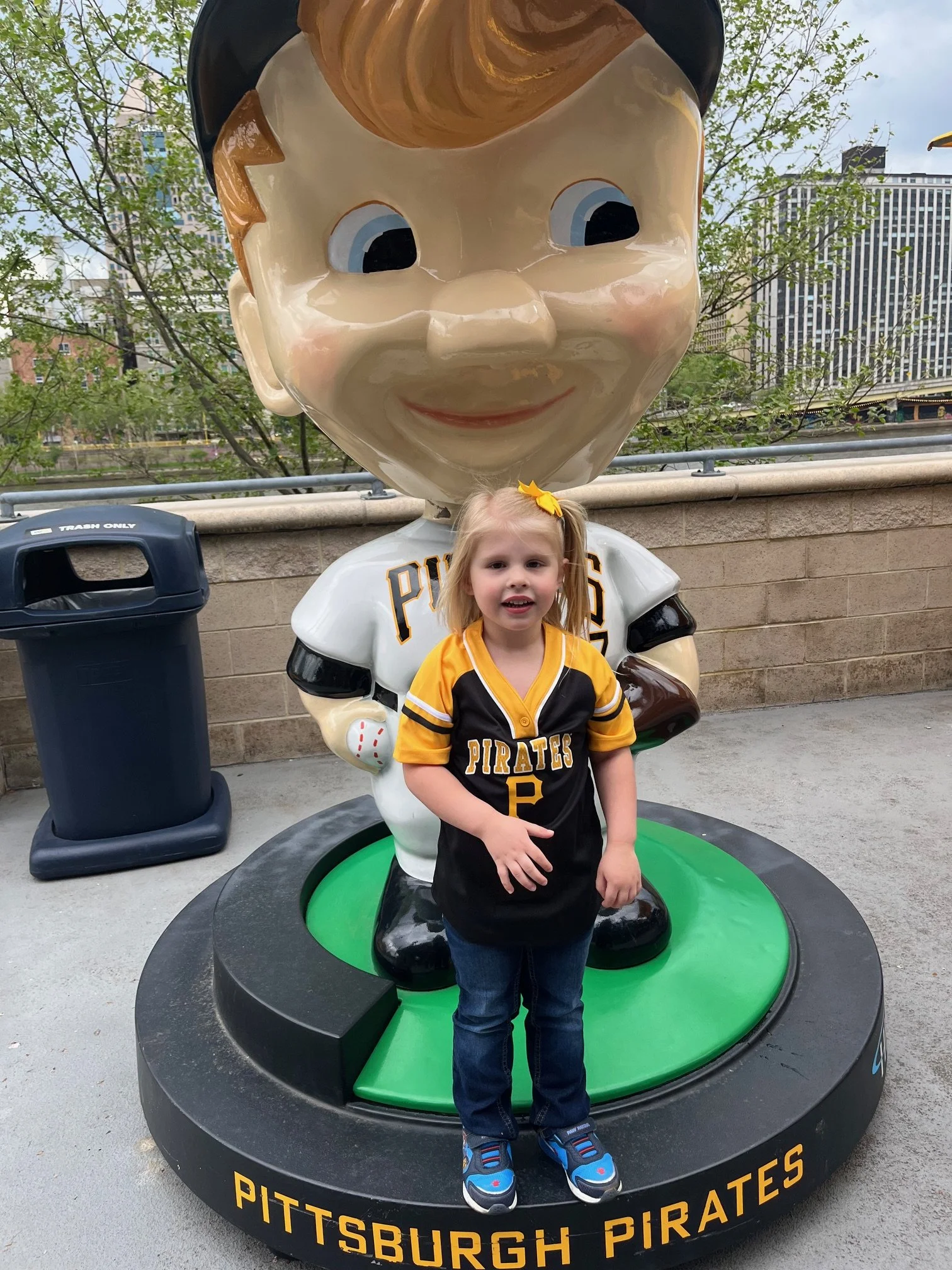 A young girl wearing a Pittsburgh Pirates baseball jersey standing in front of a large mascot statue of a smiling pirate head.