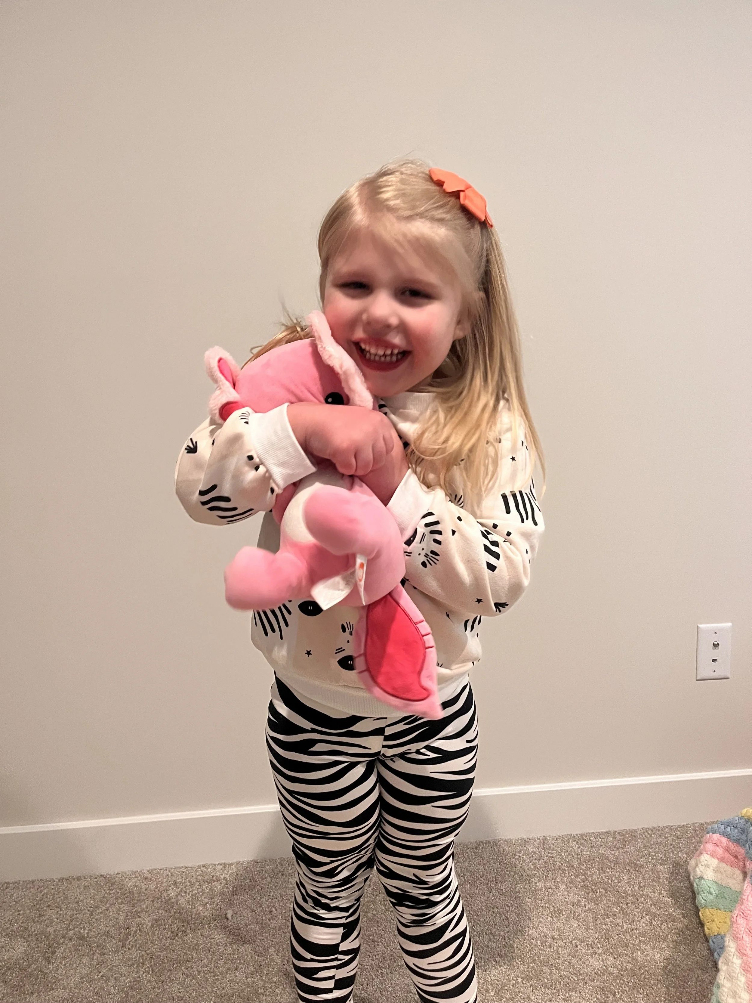 A young girl with blonde hair, wearing a white sweater with black illustrations and zebra-print leggings, is smiling and hugging a pink plush animal.