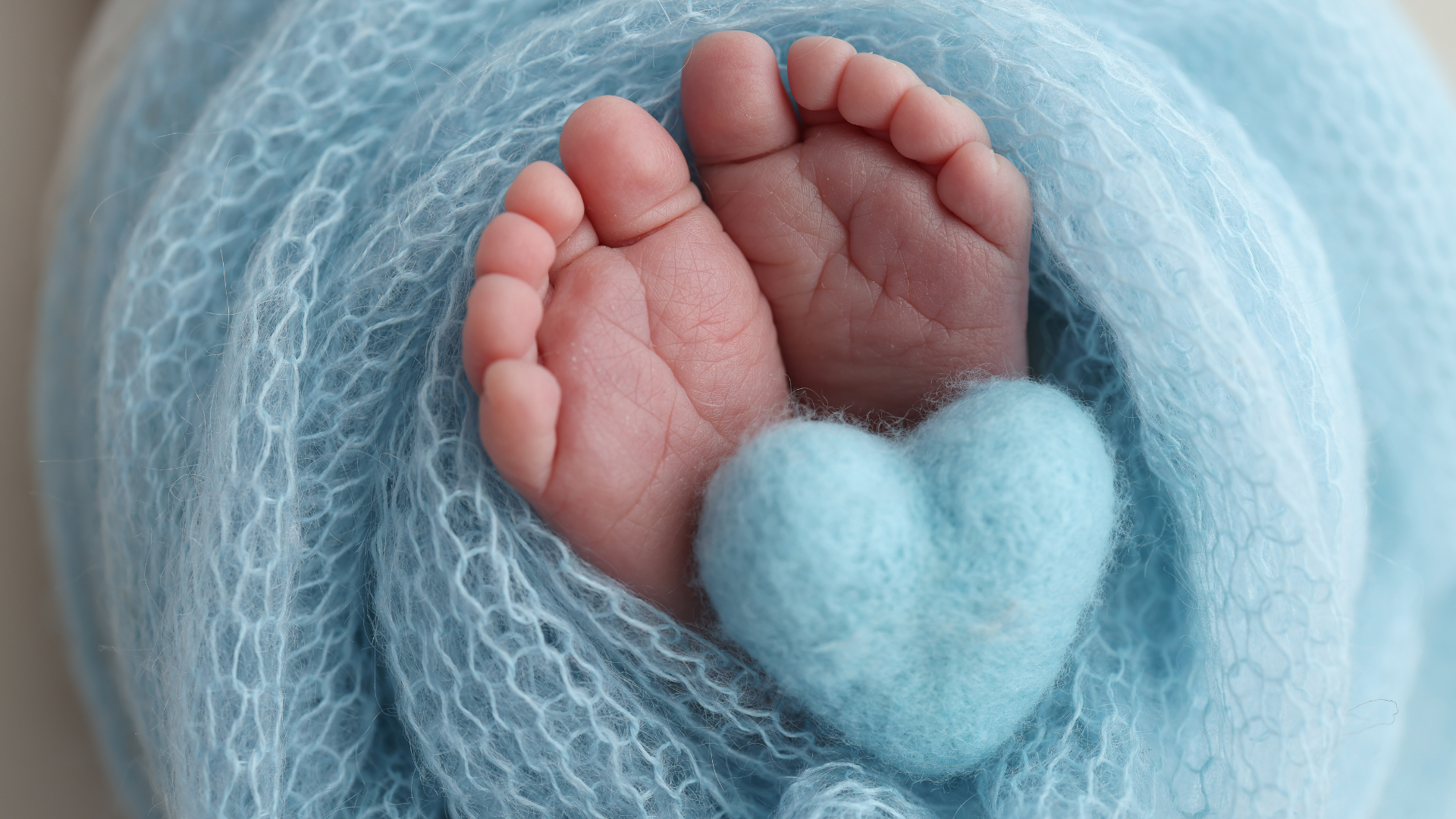 Close-up of tiny baby feet with toes curled, wrapped in soft blue fabric, with a small felt blue heart.