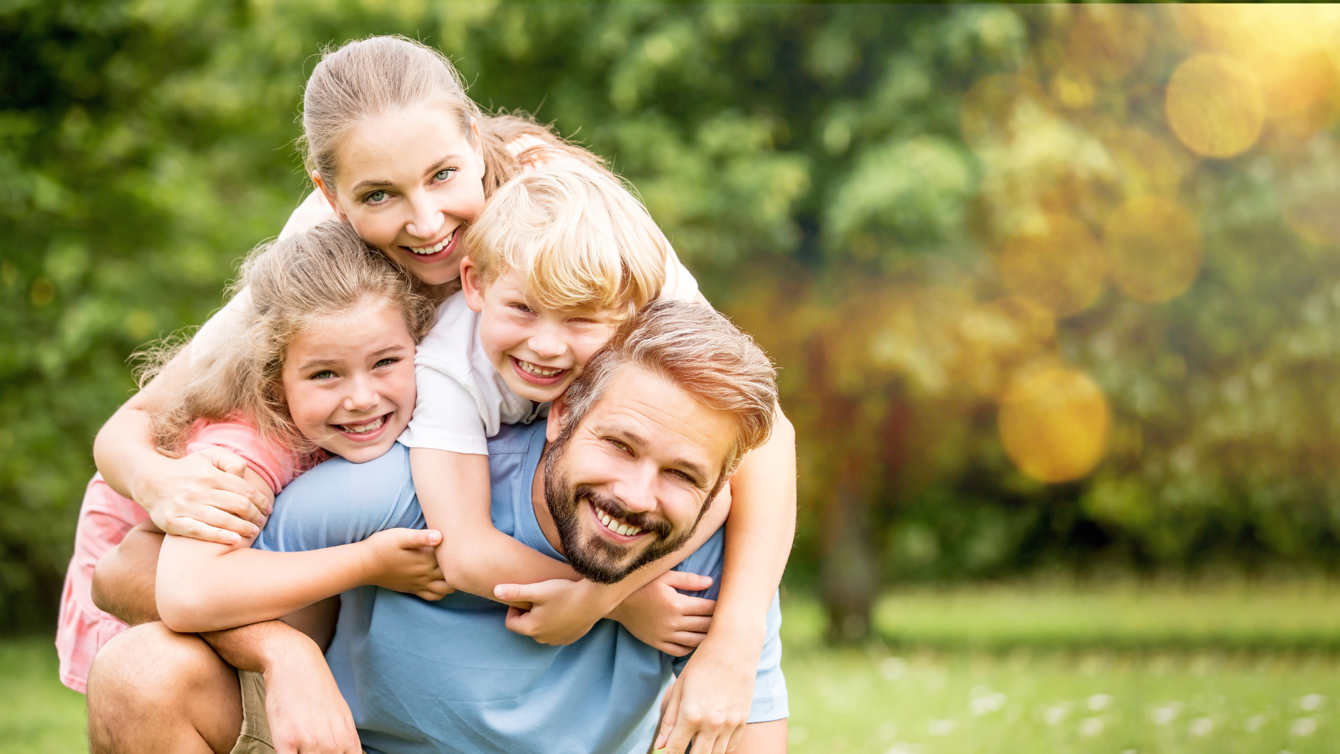 Happy family of five, with two adult parents and three children, all smiling and hugging outdoors in a park during daylight.