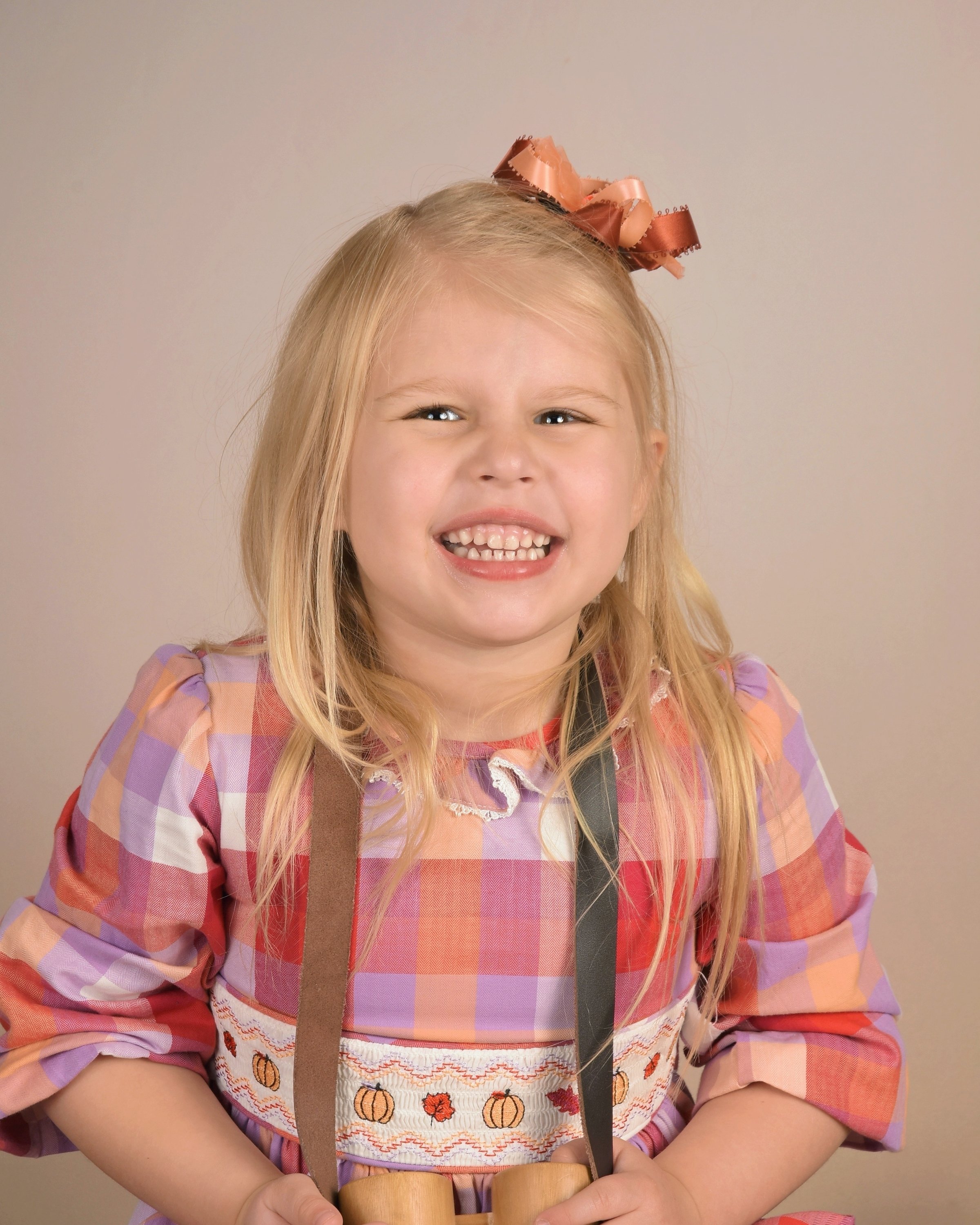 A young girl with playing jump rope, smiling, wearing a plaid dress with autumn motifs, a large bow in her hair, and holding jump rope handles.