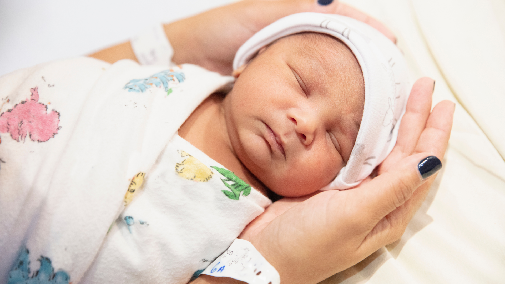A newborn baby with closed eyes sleeping, wrapped in a colorful hospital blanket, resting on a person's hand with a hospital wristband, and wearing a white hat.