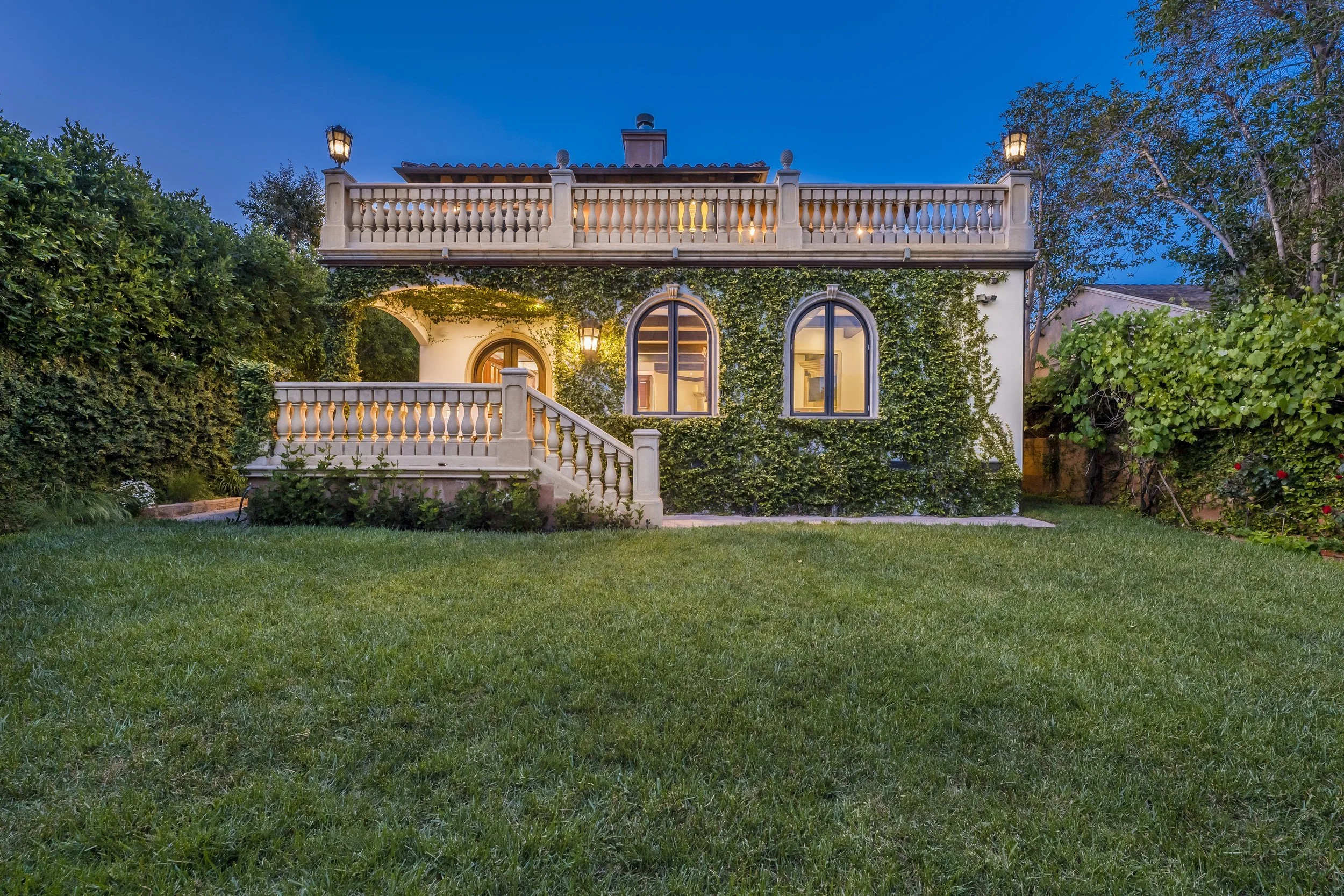 A two-story house with a balcony, illuminated by outdoor lights, surrounded by trees and a well-manicured lawn at dusk.
