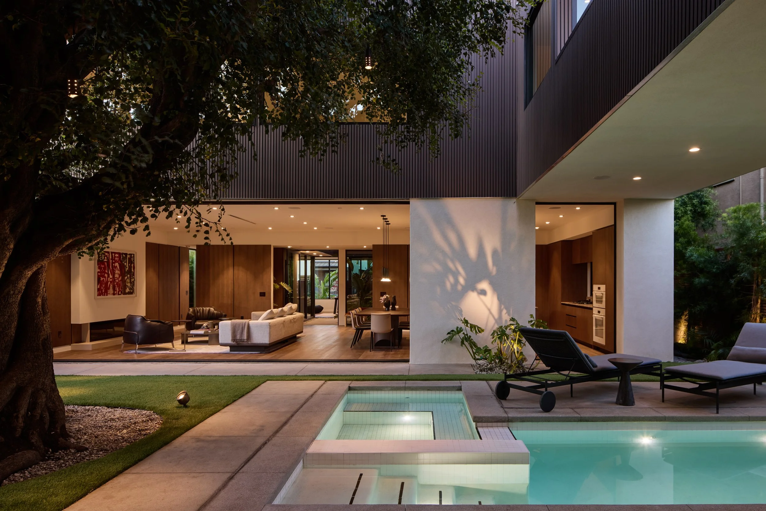 Modern house with large open living area, illuminated by ceiling lights, featuring a view of a backyard with a swimming pool, patio, and outdoor seating, surrounded by green trees and landscaped yard.