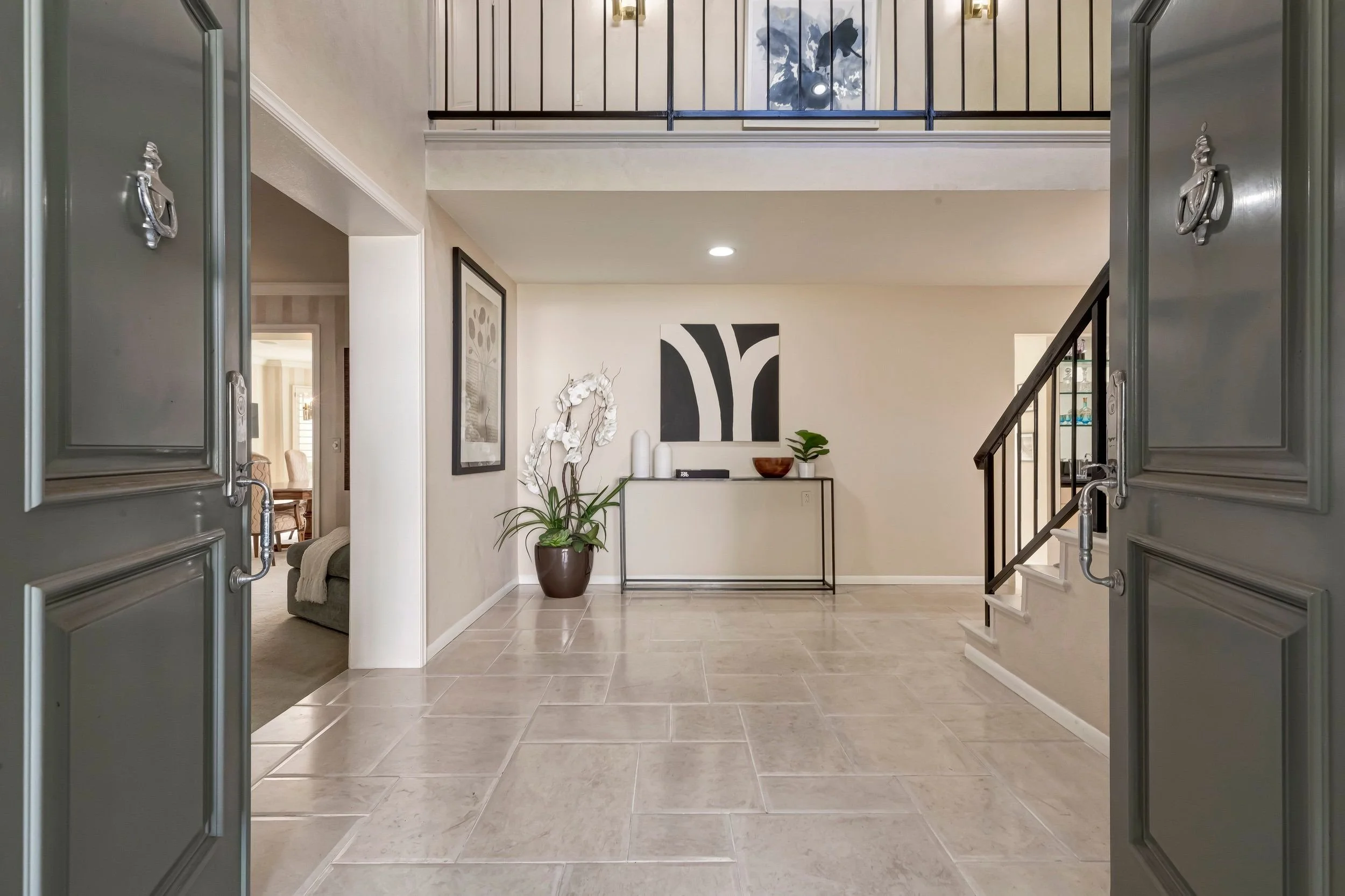 View of a home's interior entryway with an open doorway, beige walls, large decorative plant in a pot, modern wall art, and a staircase railing.