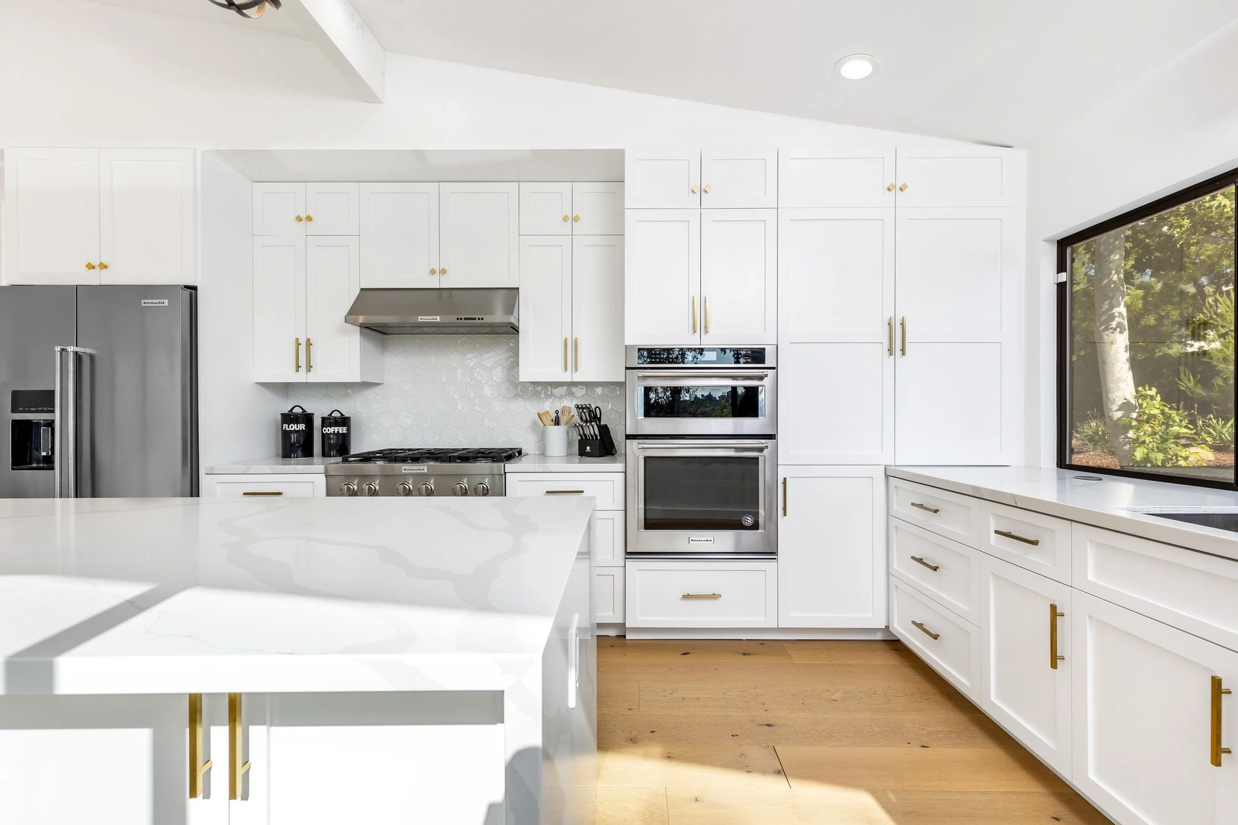 Modern white kitchen with gold hardware, stainless steel appliances, a large window, and a white countertop. Visible items include black flour and coffee containers, a white utensil holder, and a stove with a range hood.