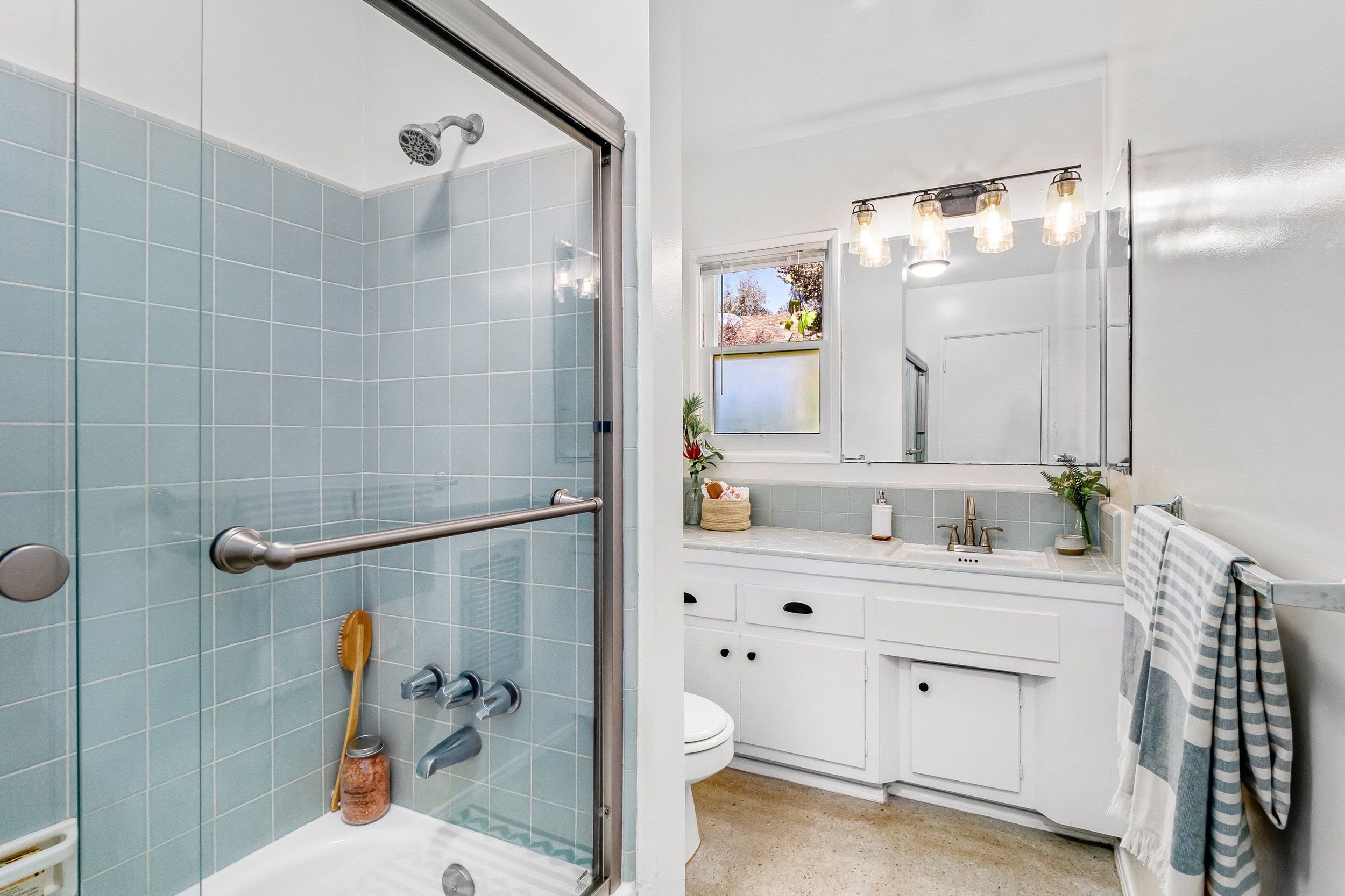 A bathroom with a walk-in shower with blue tiles, a showerhead, a glass door, a vanity with a sink, a mirror with light fixtures above, a window, and decorative plants.