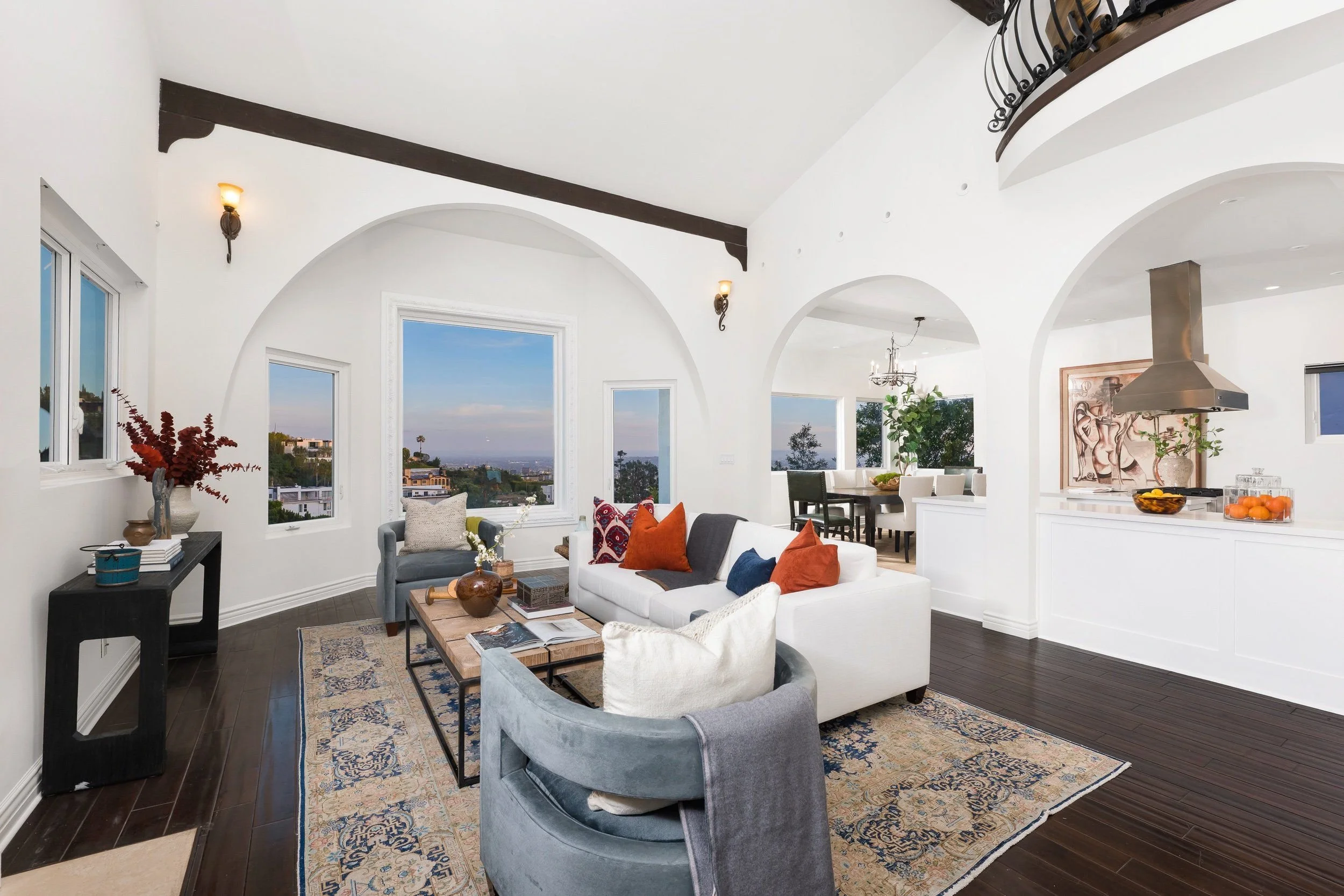 Living room with white walls, dark wooden floors, a white sofa with colorful pillows, gray armchairs, a wooden coffee table, and large windows showing an outdoor view of buildings and sky.