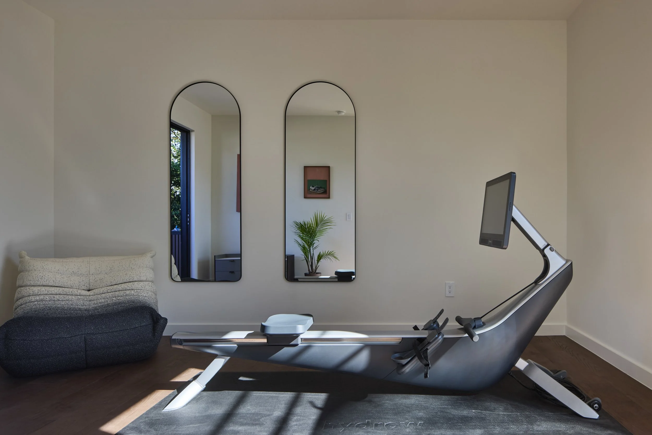 Modern living room with two tall, narrow mirrors on the wall, a potted plant, and part of a staircase. In the foreground, there's a treadmill facing the mirrors, and on the left, a textured beige sofa.