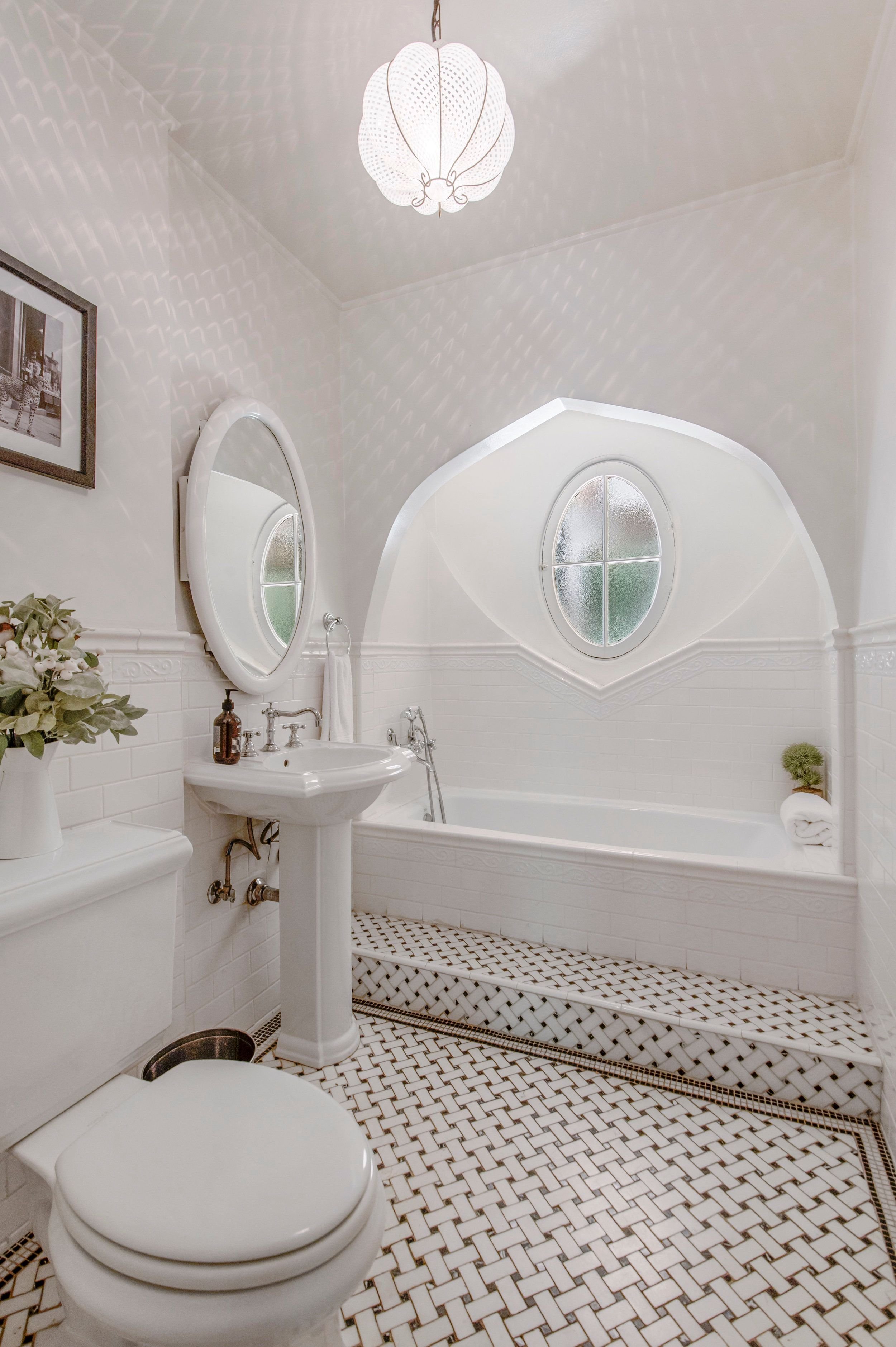 Bathroom with white tiled walls and floors, oval mirror above pedestal sink, bathtub with arched window above, black and white mosaic floor tiles, potted plant, framed photograph, and decorative lighting fixture.