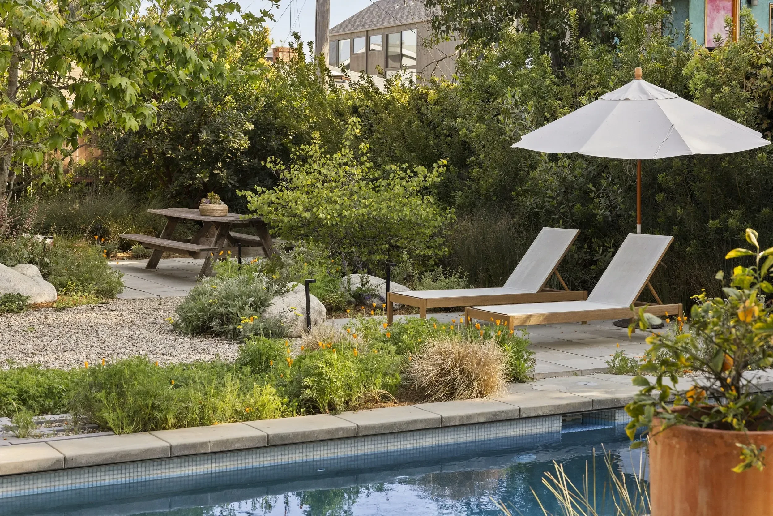 A lush backyard garden with a swimming pool, surrounded by green plants and trees, featuring a wooden picnic table with a basket of plants, a large white patio umbrella, and lounge chairs by the pool.