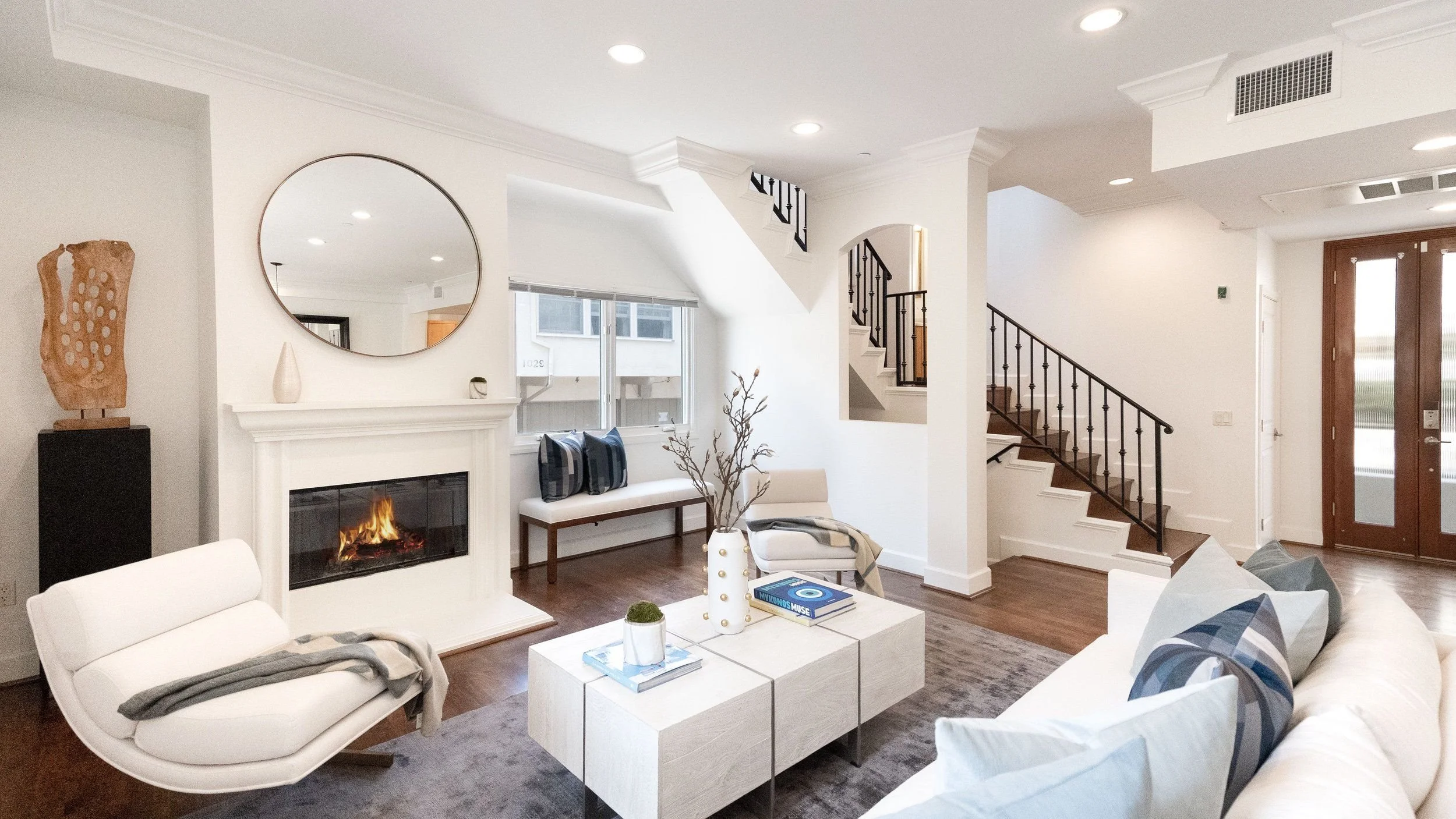 A modern living room with white walls, a fireplace, a large mirror above the mantel, a window seat with pillows, a staircase with black railing, and a front door with glass panels.
