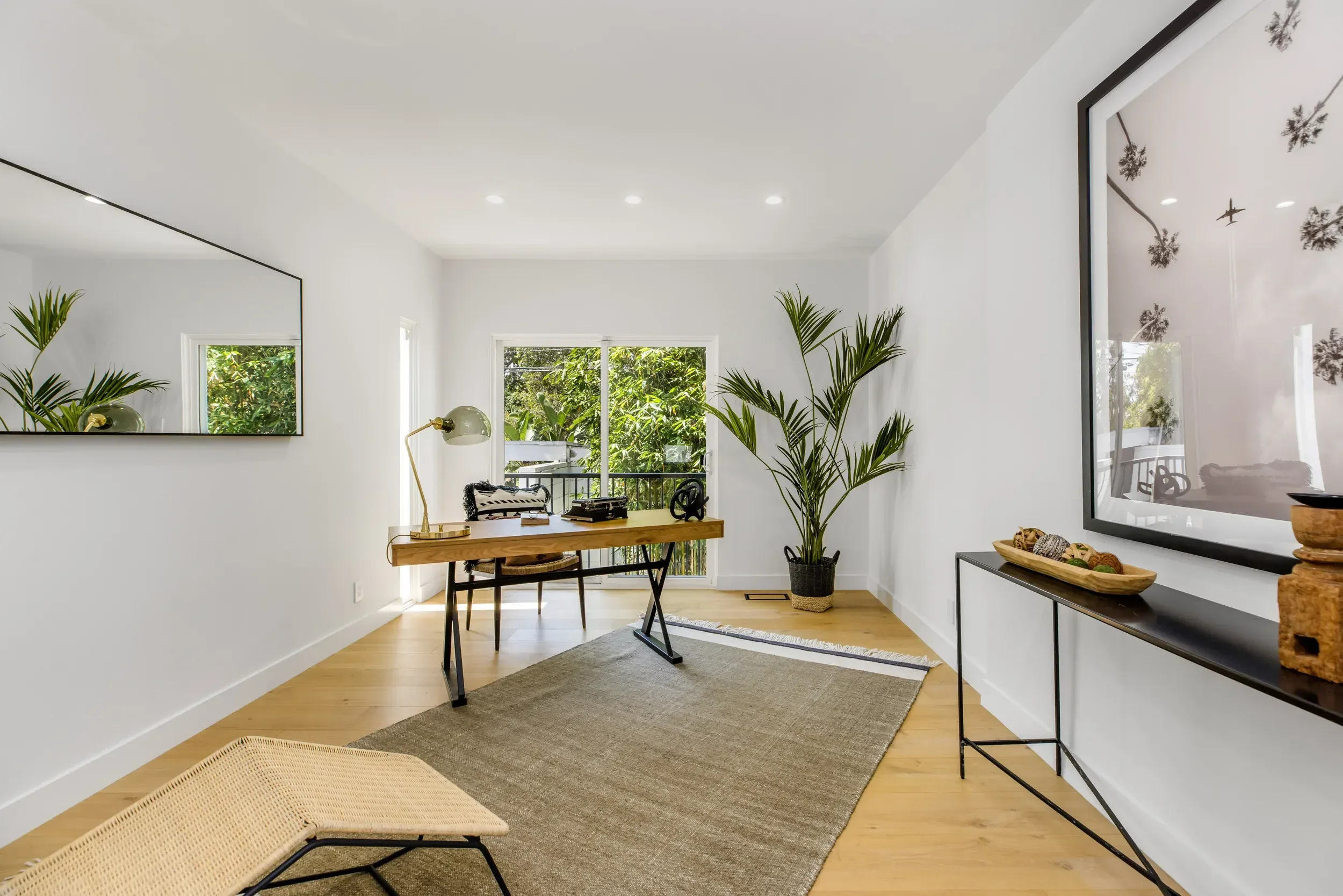 Bright and modern living room with white walls, hardwood floors, and large windows. A wooden table with chairs, a tall green potted plant, a large wall mirror, and a framed black-and-white art print are visible.