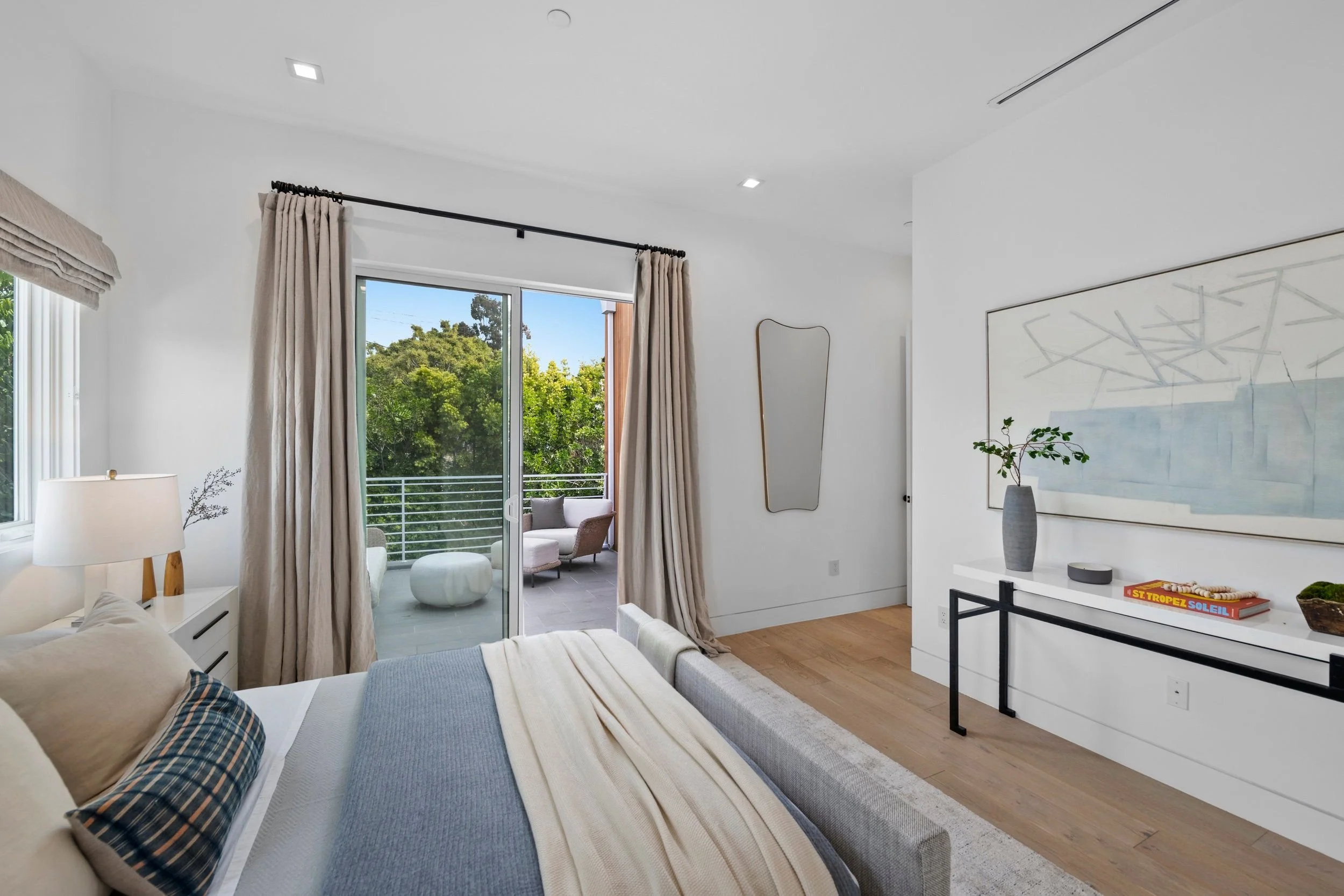 Living room with a beige sofa, brown plaid pillows, white side table, table lamp, and a balcony with outdoor seating and trees view.