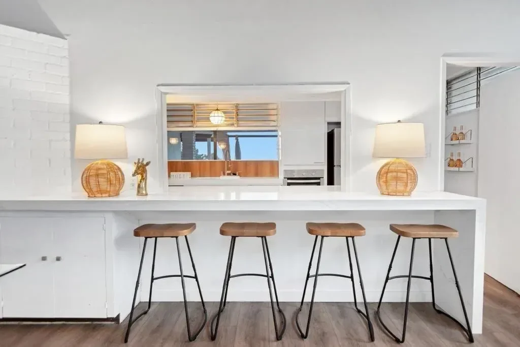Modern kitchen with white walls, four wooden bar stools at a white counter, and two table lamps with rattan bases on the counter. Through the opening, a kitchen with a wooden backsplash and a window view of trees is visible.