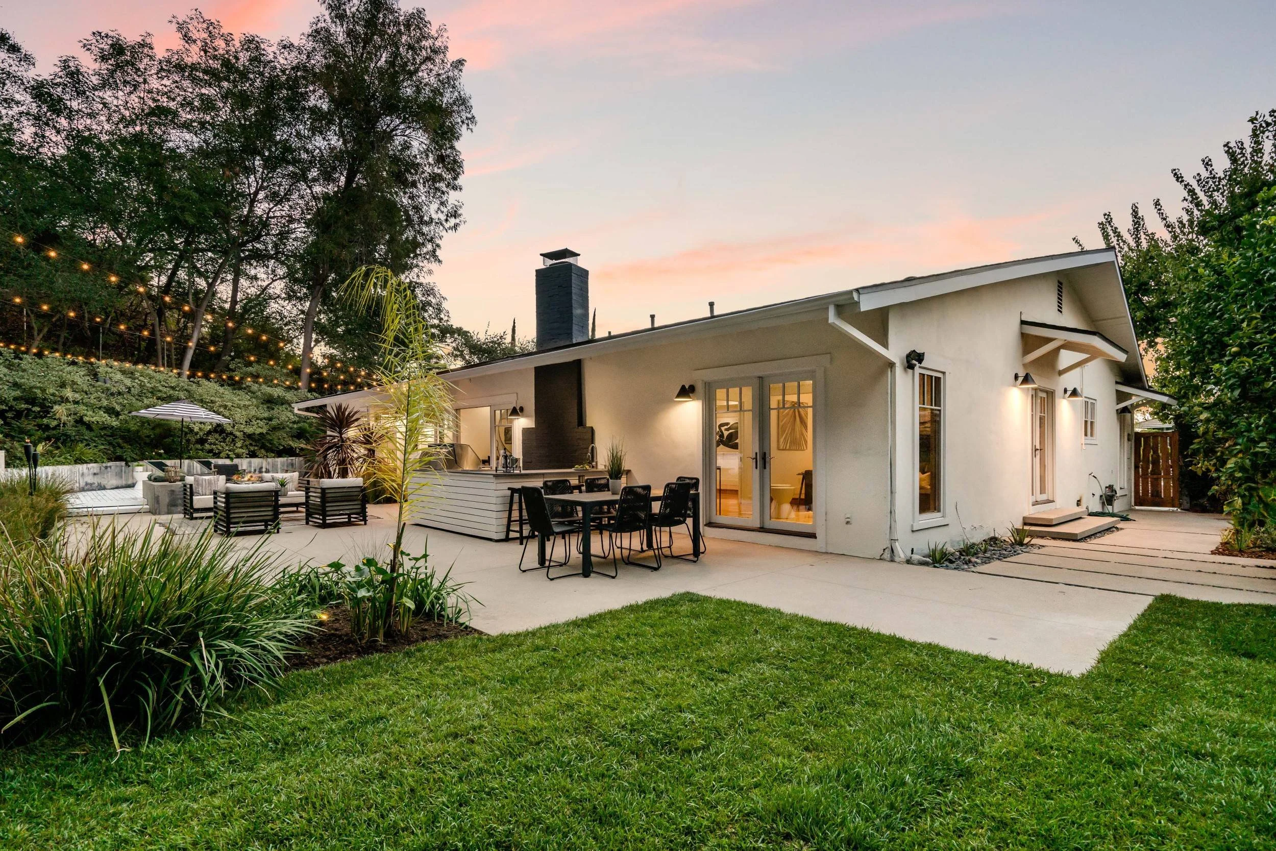 A backyard with a modern house, outdoor dining area, and string lights at sunset.