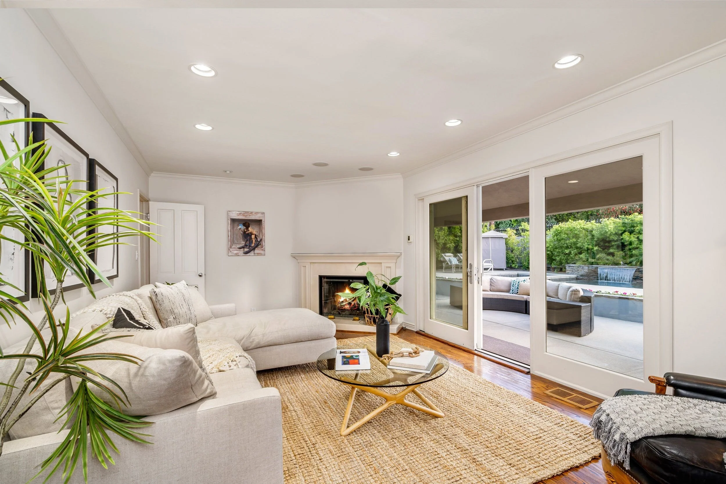 A bright living room with white walls, a fireplace, and large sliding glass doors leading to an outdoor patio. There are beige sofas, a round coffee table, indoor plants, and framed artwork on the walls. The room has wooden flooring and a woven rug.