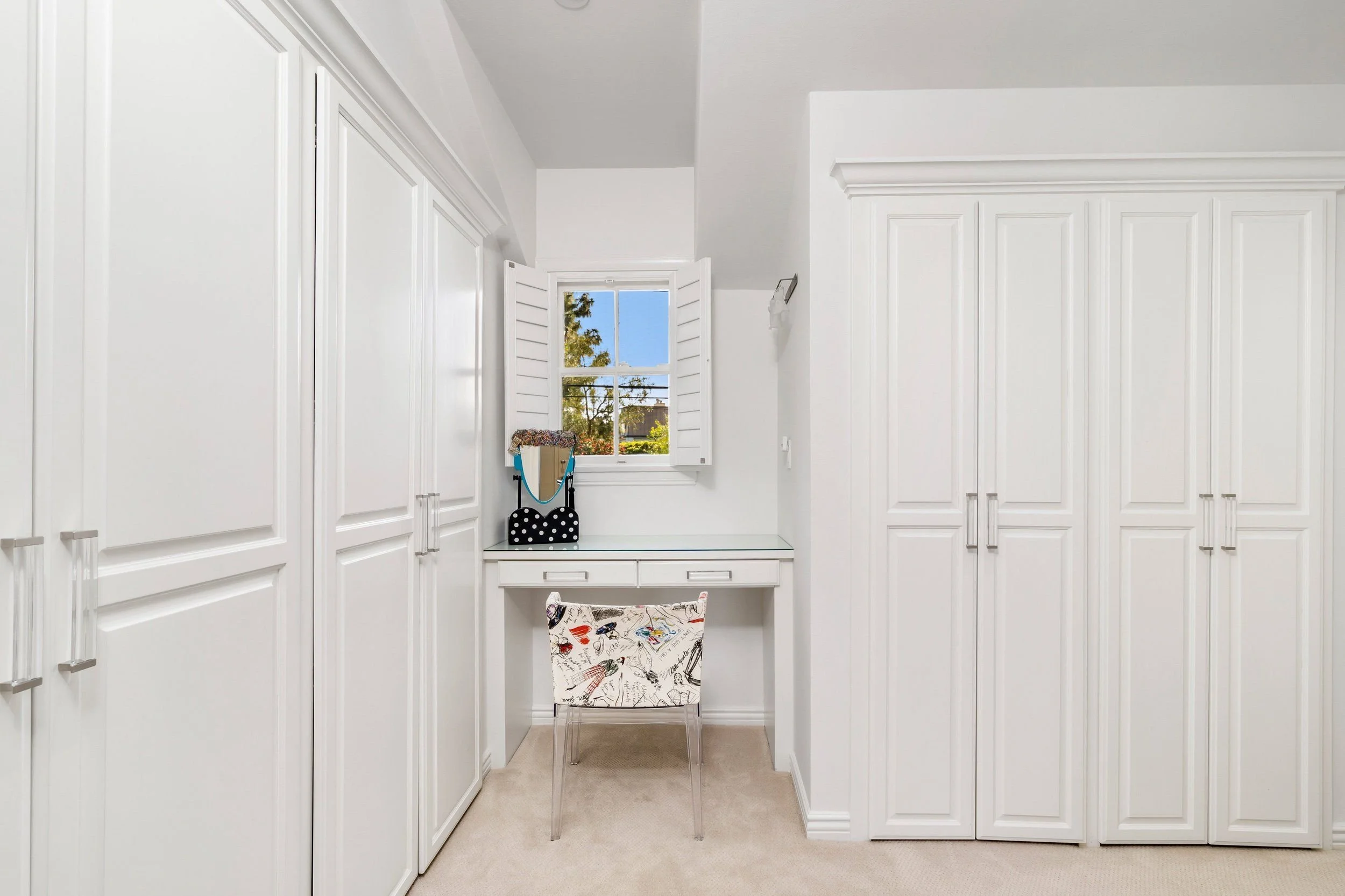 A walk-in closet with white built-in cabinets on both sides, a small window with shutters, a mirrored table with a black and white polka dot box, and a chair with a colorful cushion, with carpeted flooring.