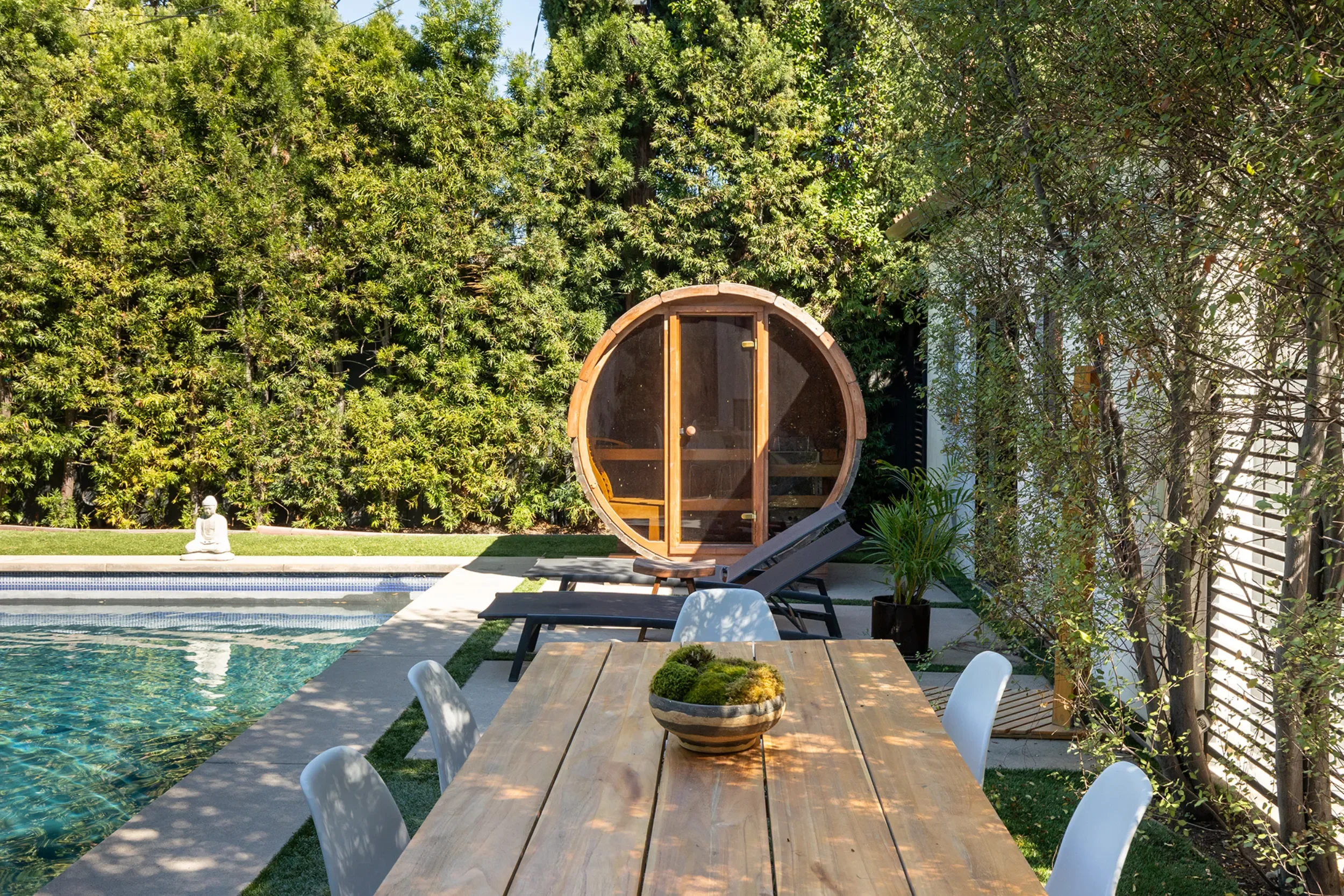 A backyard with a swimming pool, wooden hot tub, outdoor dining table with chairs, lounge chairs, a small Buddha statue, lush green trees, and a partially visible house.