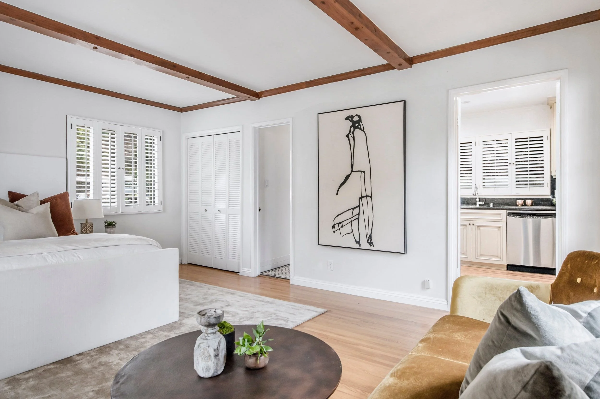 A living room with white walls, wooden beams on the ceiling, a large window with plantation shutters, a white bed with pillows, a round coffee table with vases and plants, and a framed abstract art piece on the wall.