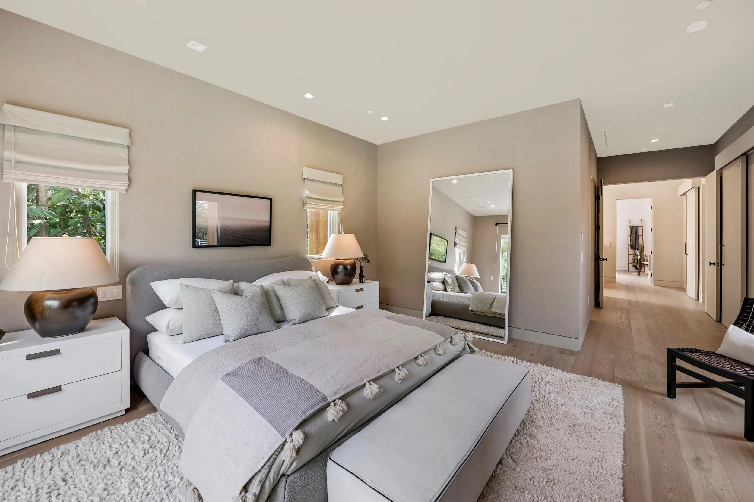 A modern bedroom with a grey upholstered bed, white nightstands, beige walls, three windows with white roman shades, a large mirror, a black and white framed picture, and a wooden floor with a fluffy white rug.