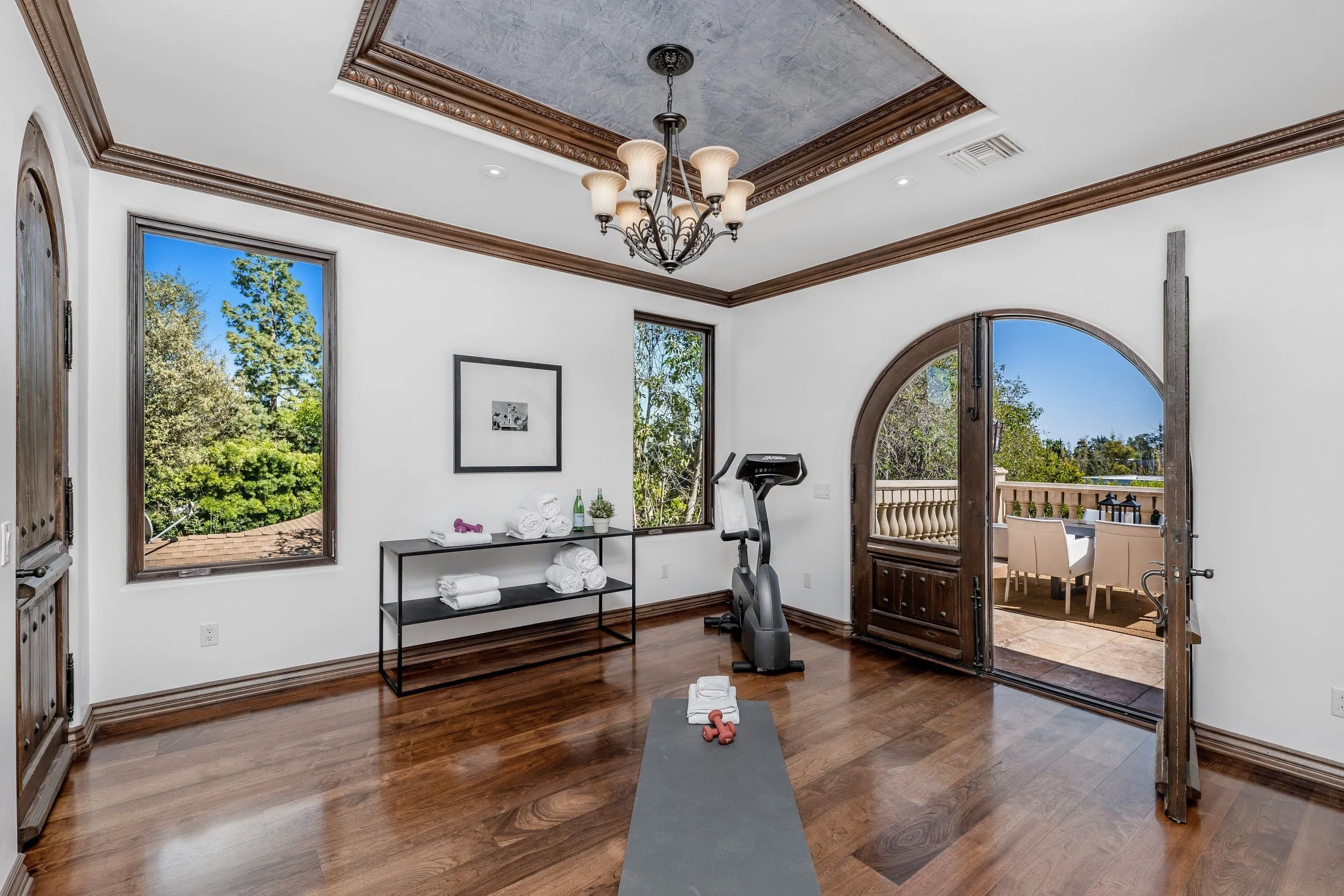 Bright exercise room with hardwood floors, large windows showing trees outside, a ceiling light fixture with multiple lamps, and a treadmill against the wall. There is a black shelving unit with rolled towels, bottles, and a plant, as well as a yoga 