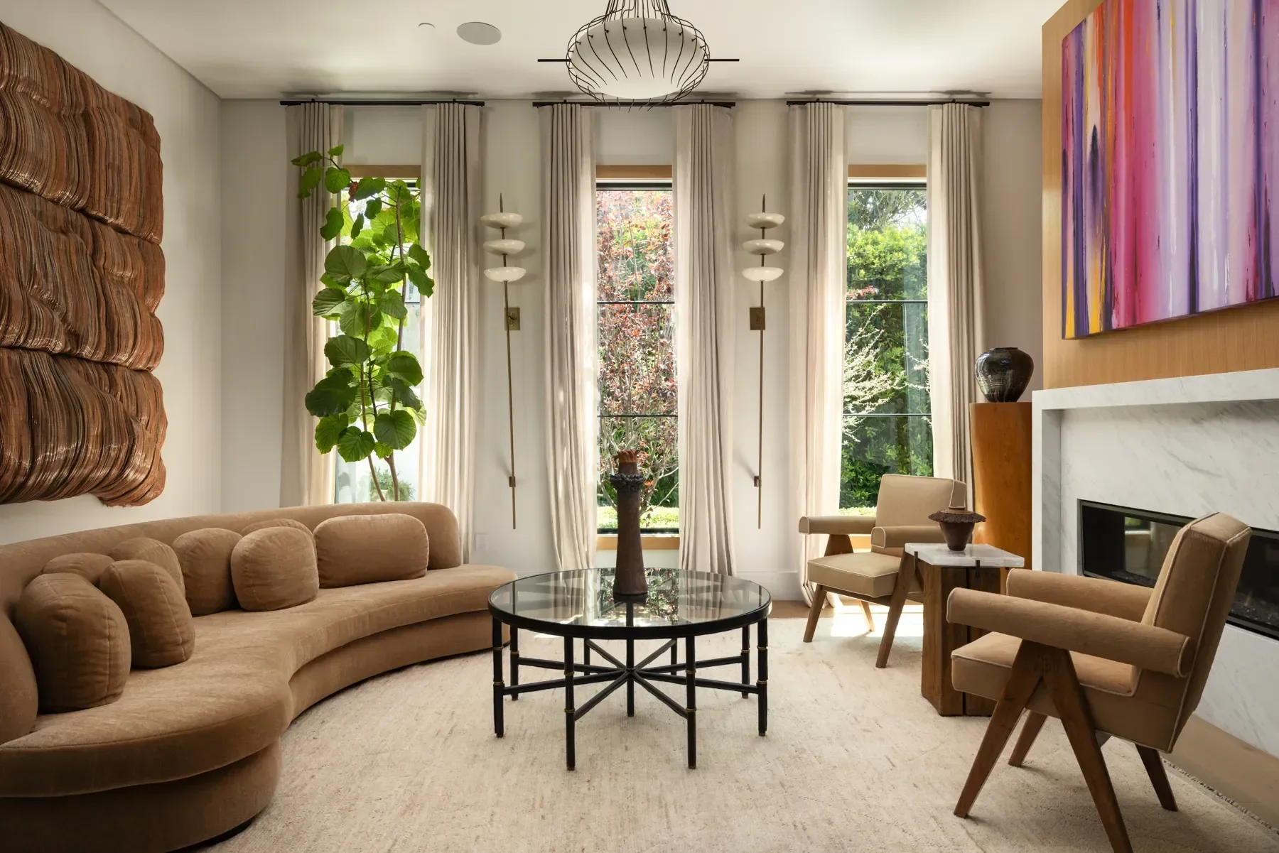 Living room with beige sofa, black round coffee table, beige armchair, white curtains, large windows with greenery outside, colorful abstract painting above fireplace, potted plant, decorative lamps, and neutral-toned decor.