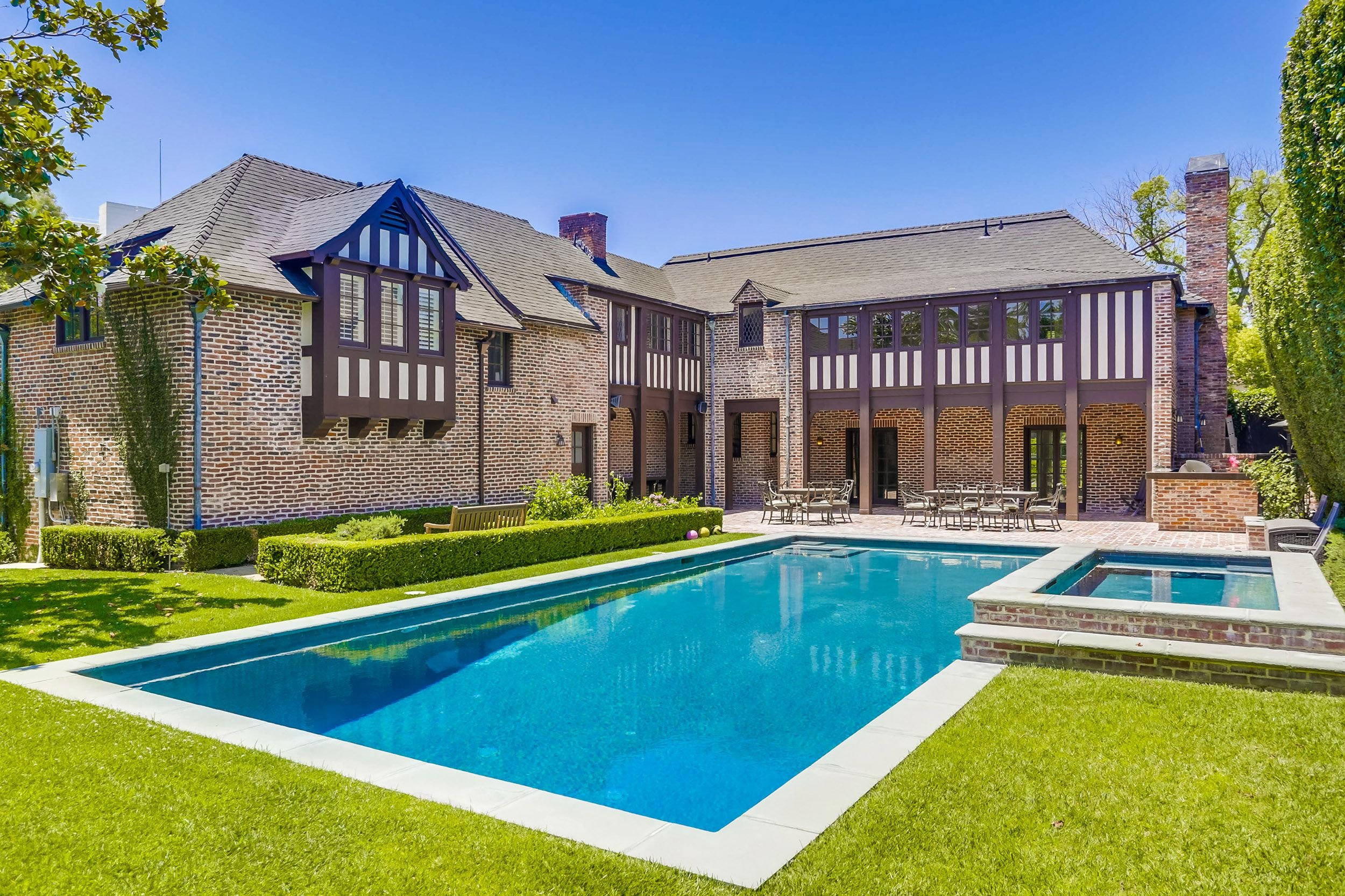 Backyard view of a brick house featuring a swimming pool, patio furniture, and green lawn under a clear blue sky with trees.