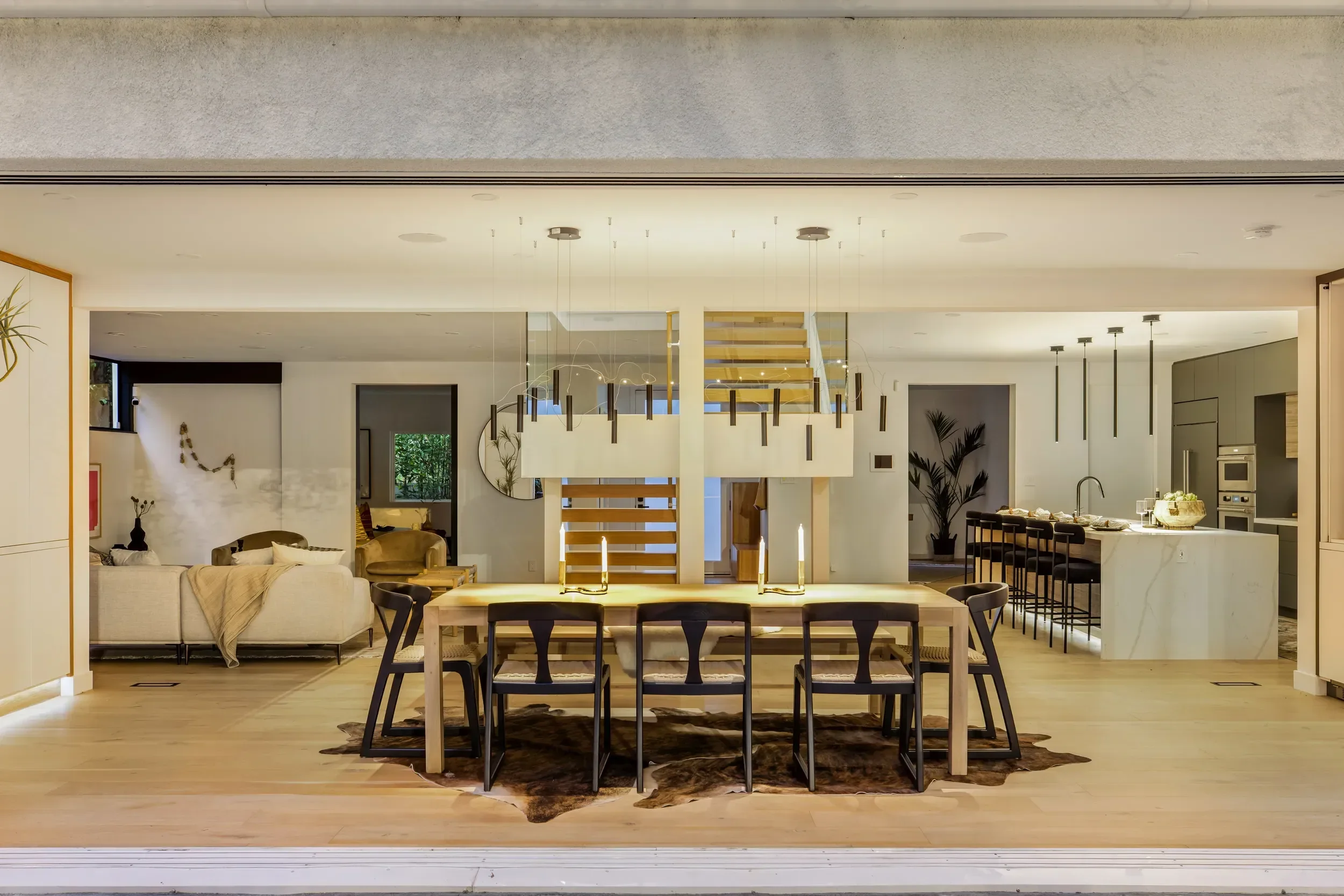 Open-concept living room and kitchen with modern decor, including a dining table with candles, beige sofa, bar stools at kitchen island, and decorative plants.