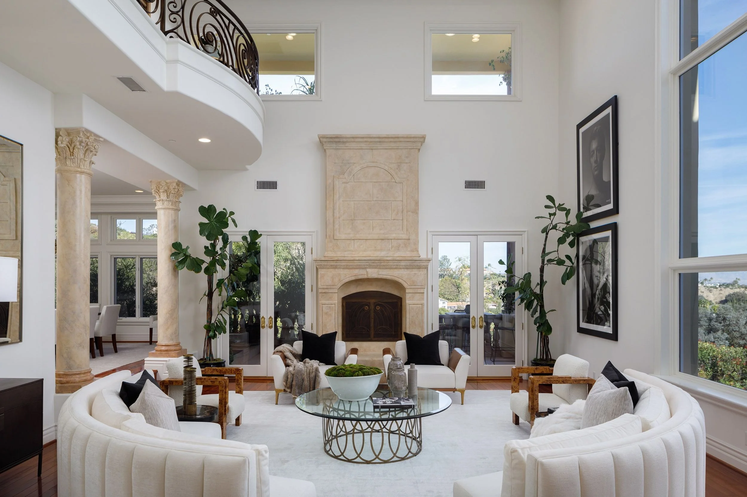 Bright living room with white curved sofas, a glass coffee table, and large windows showing a scenic view. There are tall plants and black and white framed photographs on the wall. The room features cream-colored columns and a fireplace with a beige 