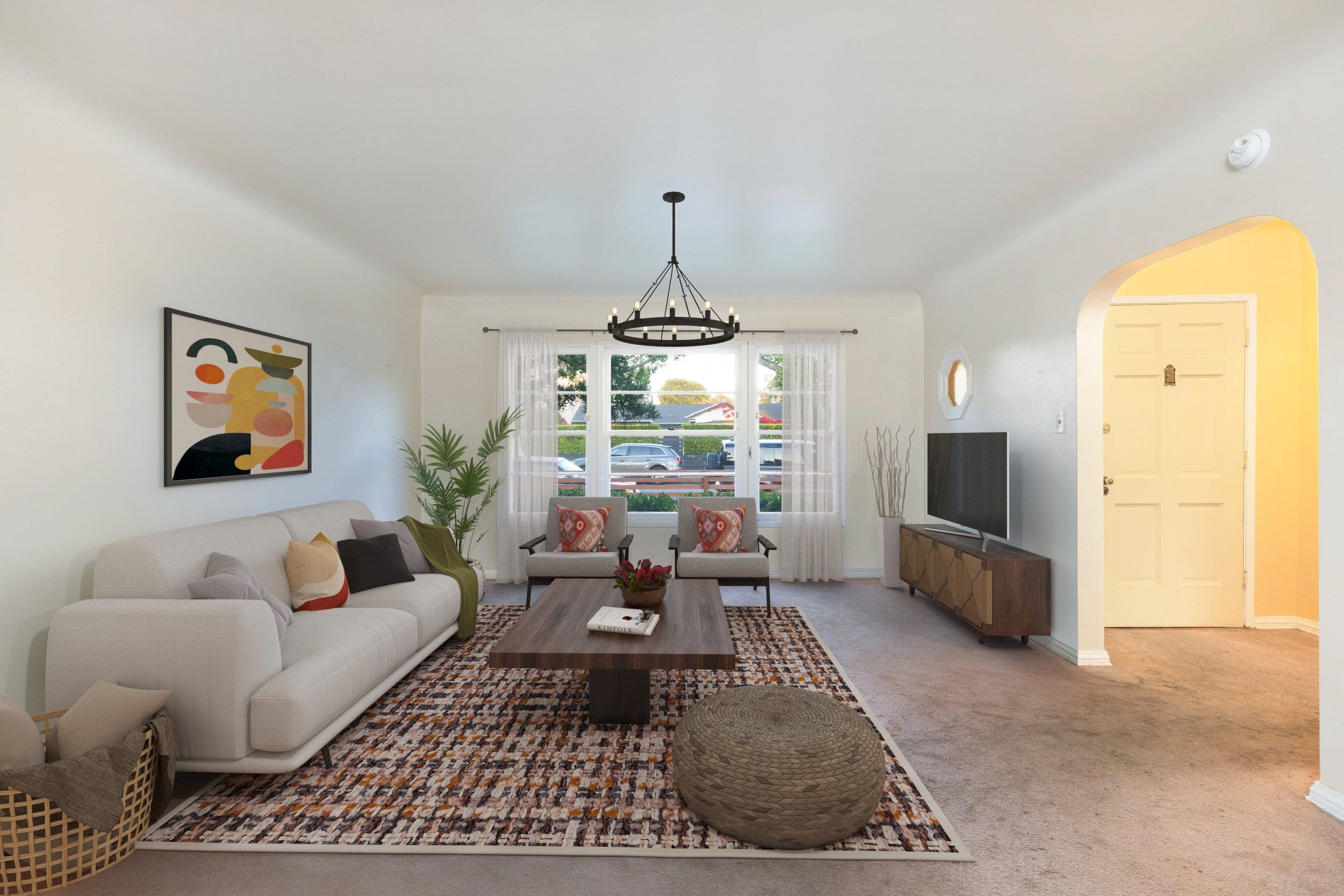 Living room with white sofa, armchairs with patterned cushions, wooden coffee table, textured ottoman, colorful abstract wall art, large window with sheer white curtains, potted plant, ceiling chandelier, TV on a wooden stand, and an archway leading 