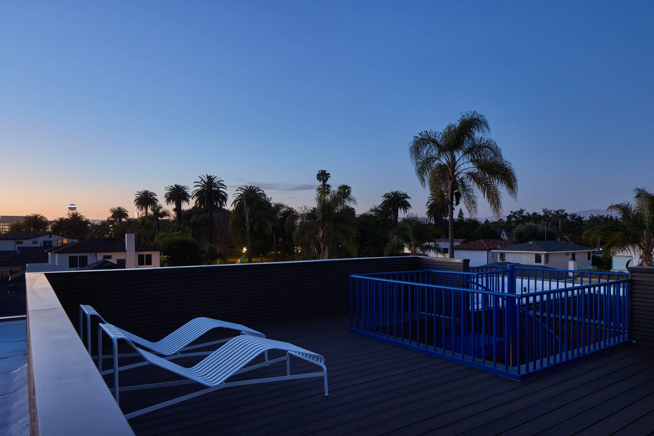 View of a rooftop deck with white lounge chairs, a blue safety railing, and neighboring houses, with tall palm trees and a clear blue sky at dusk.