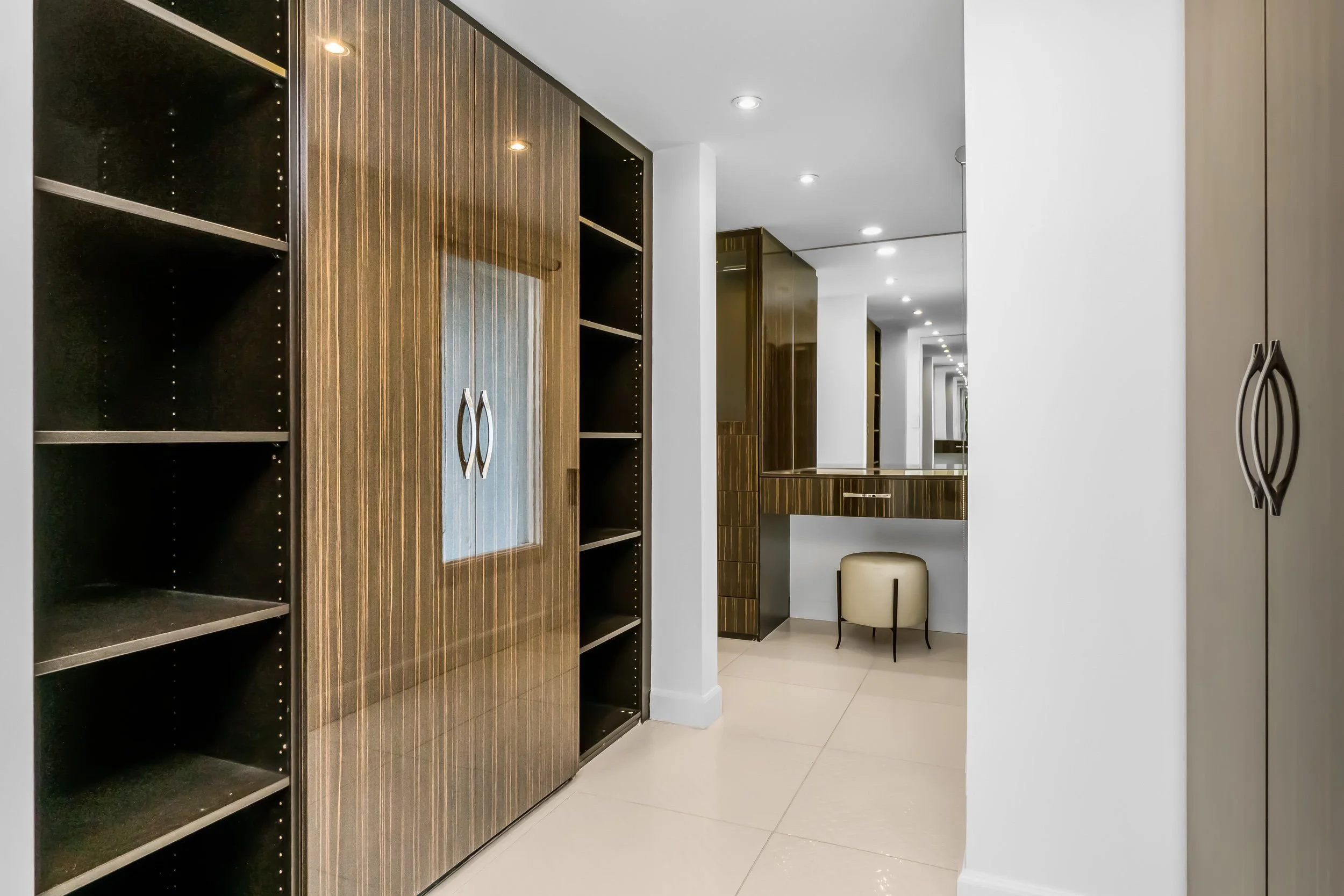 Clean modern hallway with wooden closets, white walls, mirrored surface, and beige tile flooring, with a small beige stool and arching chair in the background.