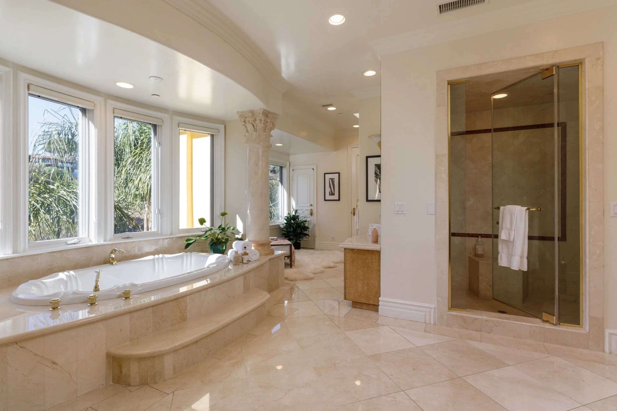 Luxurious bathroom with a large bathtub under windows, a marble shower enclosure, and elegant decor including framed art and plants.