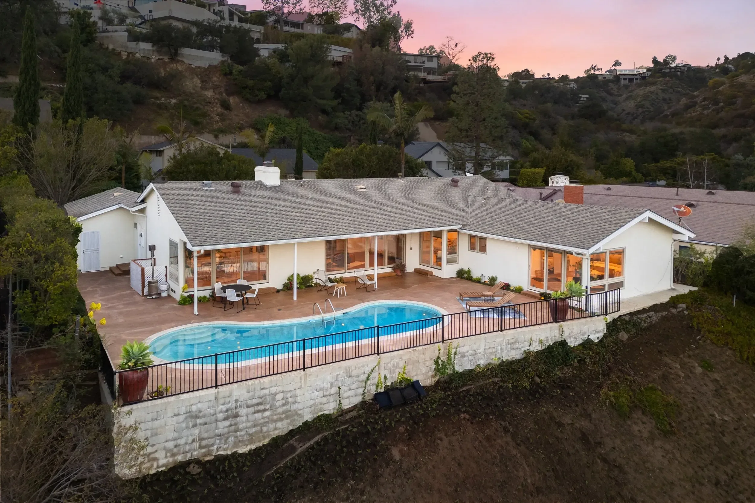 A house with a swimming pool in the backyard, surrounded by a black fence, with patio furniture and potted plants, at sunset with a view of hillside houses and trees.