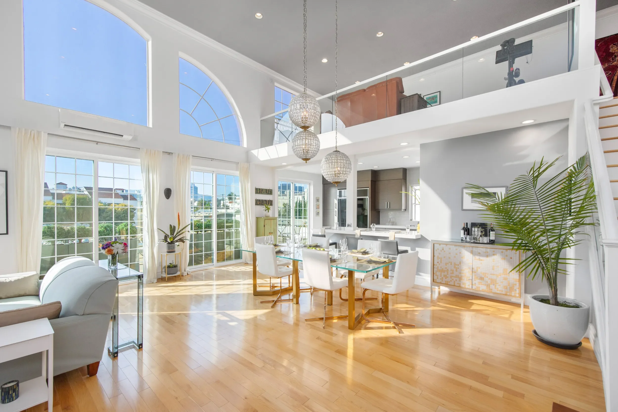 Bright, spacious living and dining area with large windows and a view of neighboring houses, featuring white furniture, plants, and modern decor.