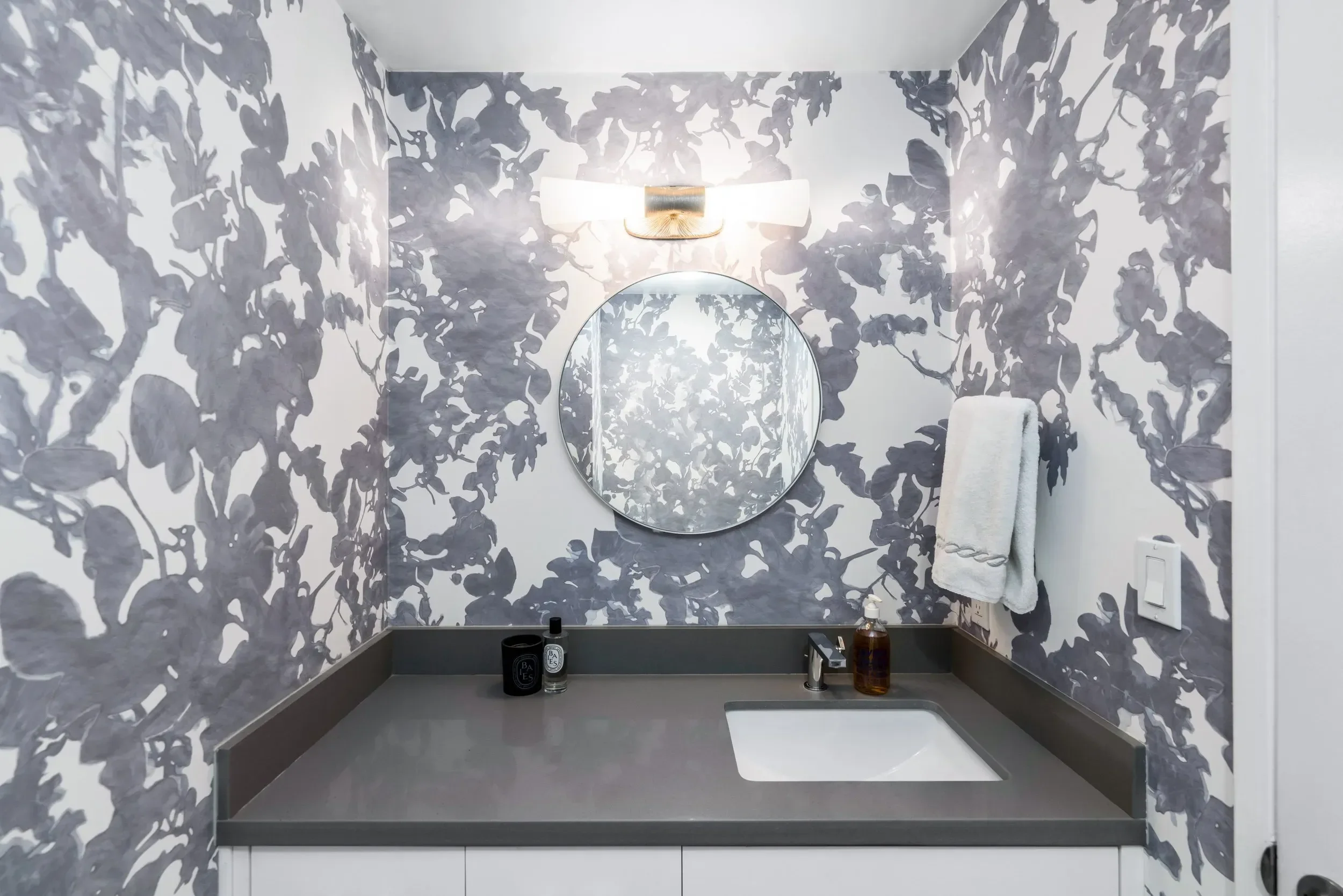A modern bathroom with gray and white floral-patterned walls, a round mirror, and a white towel hanging on a hook. On the gray countertop, there are various toiletry bottles and a brown soap dispenser.