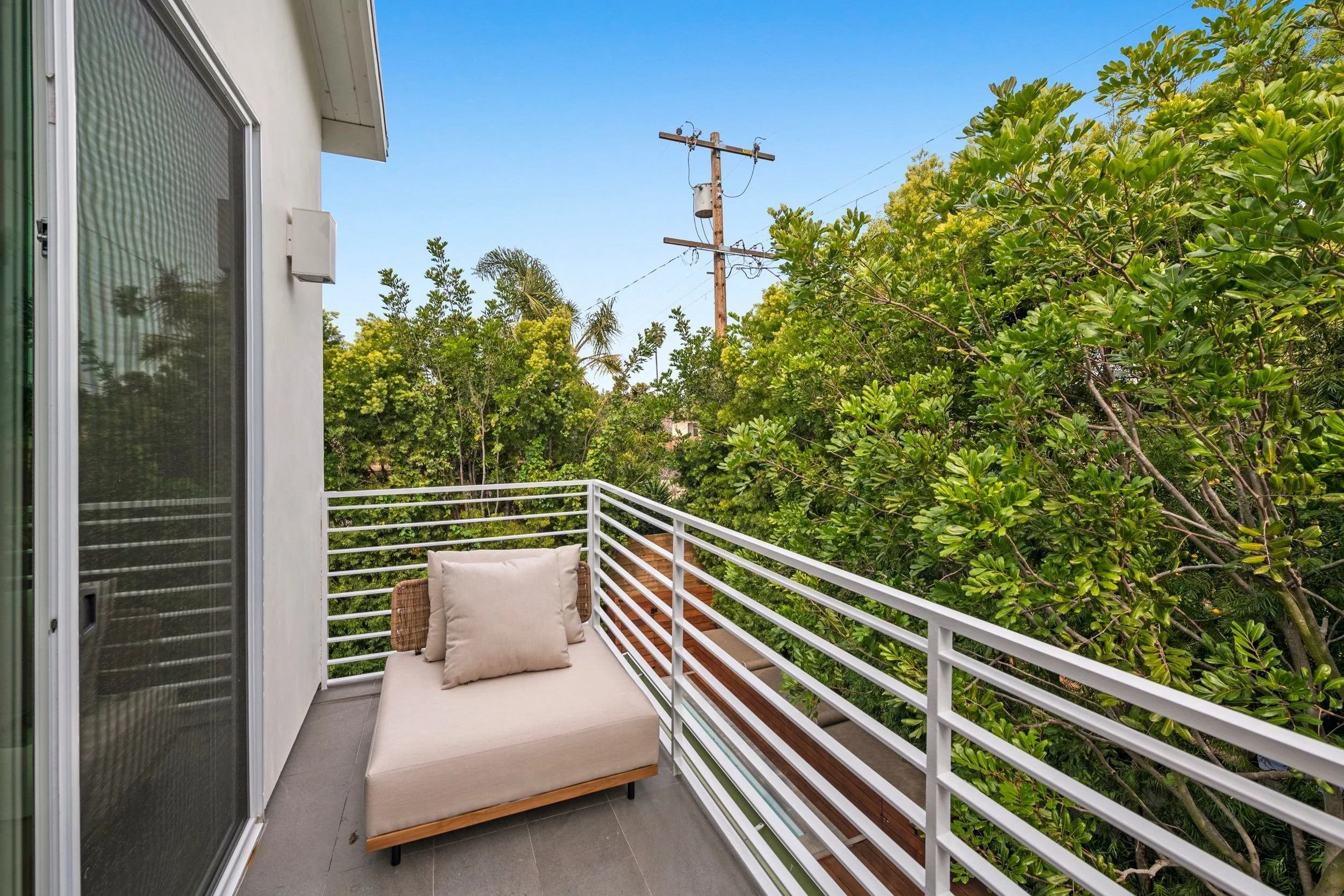 A balcony with a beige cushioned chair and pillow, white railing, green trees, and a clear blue sky.