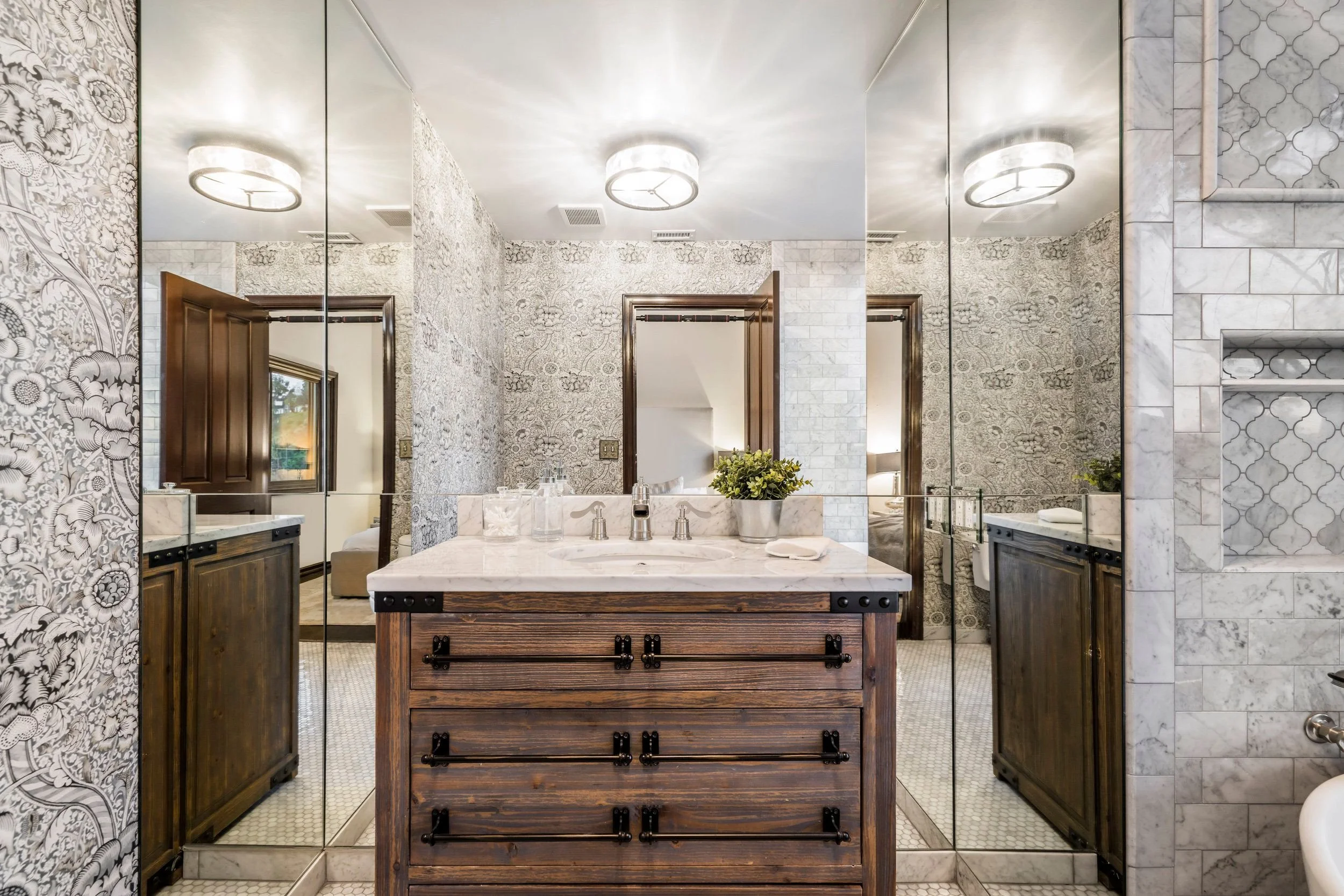 A bathroom with a large mirror above a marble countertop. The bathroom has patterned wallpaper and a wooden cabinet under the sink. There is a small potted plant and a soap dispenser on the countertop. The bathroom features a glass-enclosed shower wi