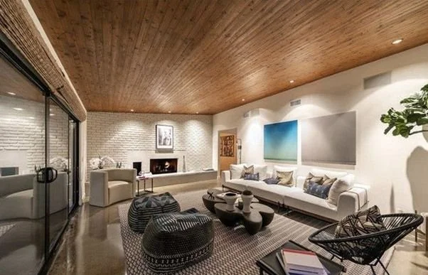 Modern living room with white sofa, black wicker ottomans, glass coffee table, wooden ceiling, and art on the white walls.